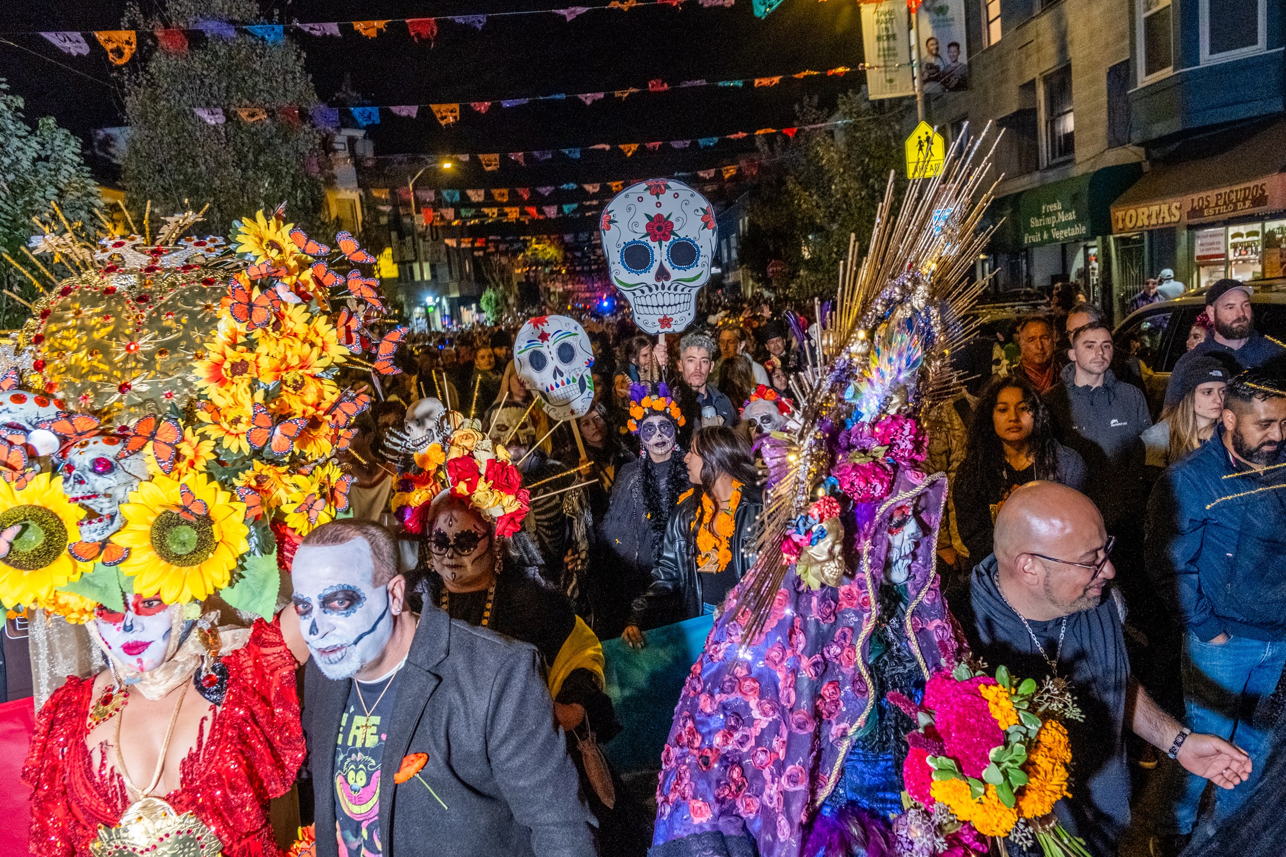 The image shows a vibrant Day of the Dead parade at night, with people dressed in colorful outfits, painted faces, and large floral headpieces, holding giant skulls.