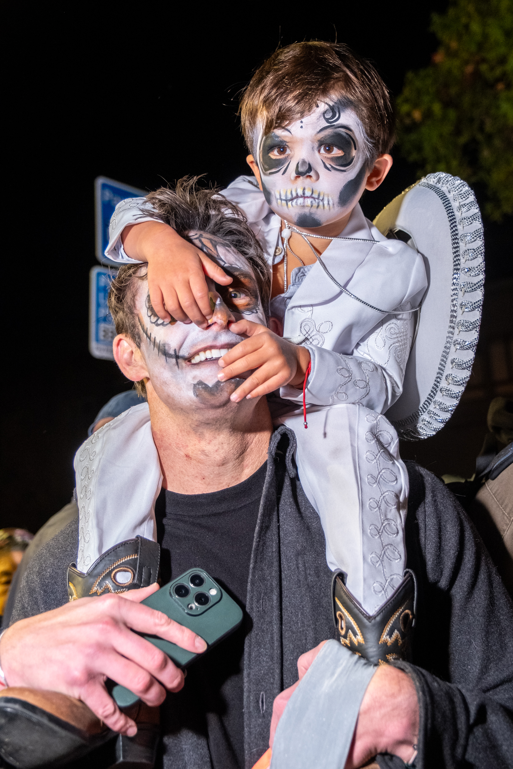 A child in skeleton face paint and mariachi costume sits on a man's shoulders, who also has painted face, holding a smartphone amidst a night scene.