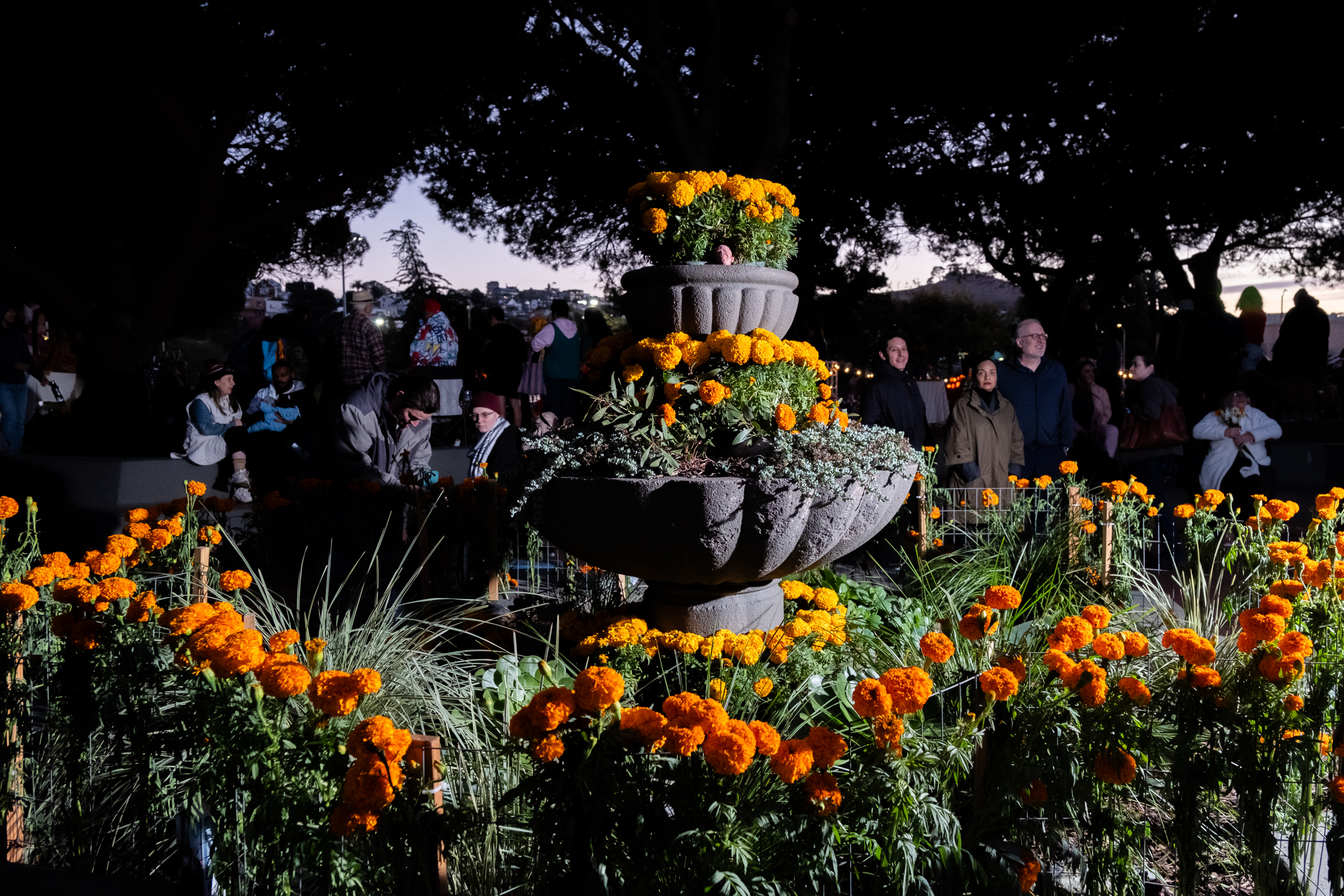 A stone fountain is adorned with vibrant orange marigolds, surrounded by people sitting and standing in the dimly lit garden setting.