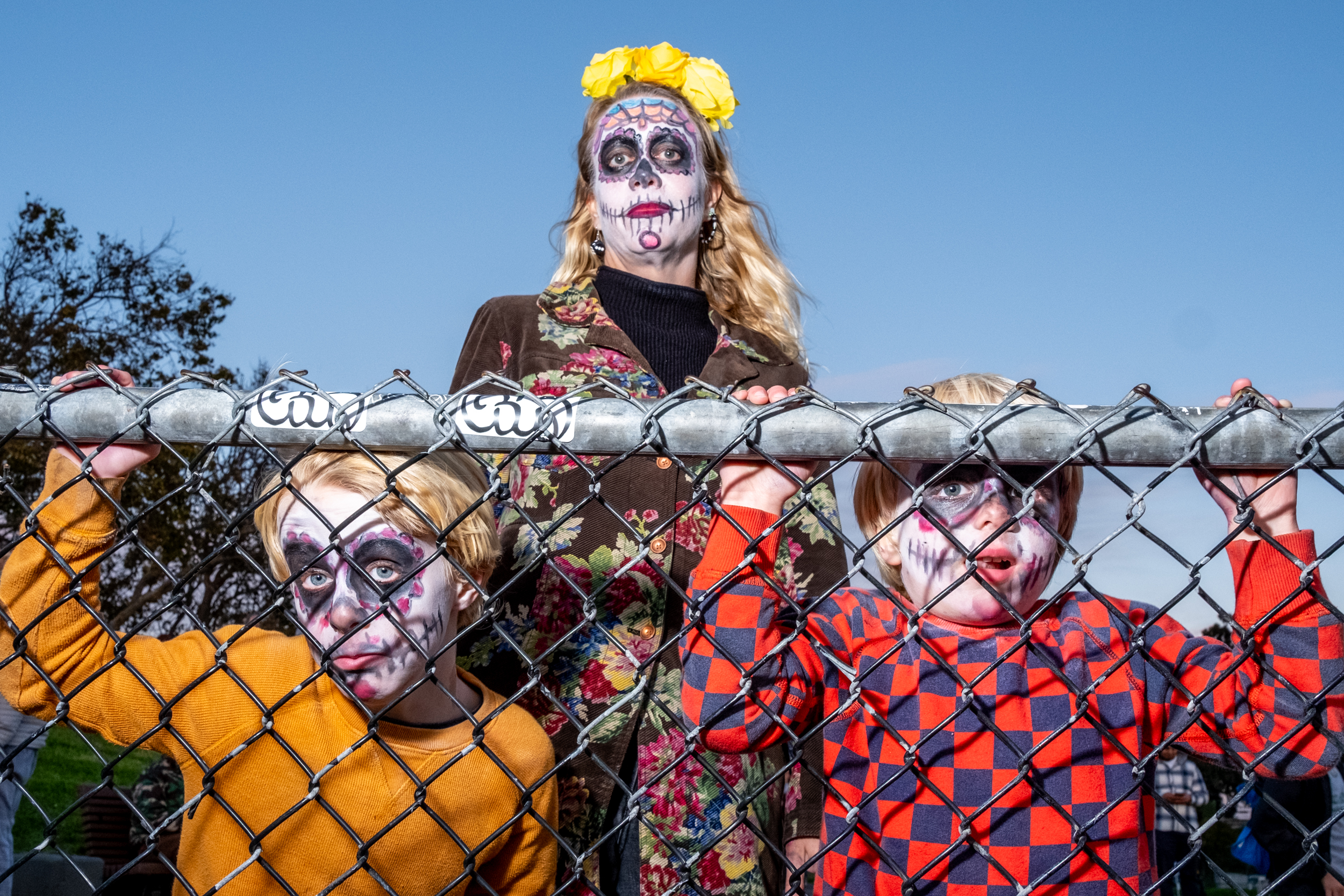 A person with sugar skull makeup and yellow flowers in their hair stands behind a fence. Two children, also with skull makeup, peek through the fence.