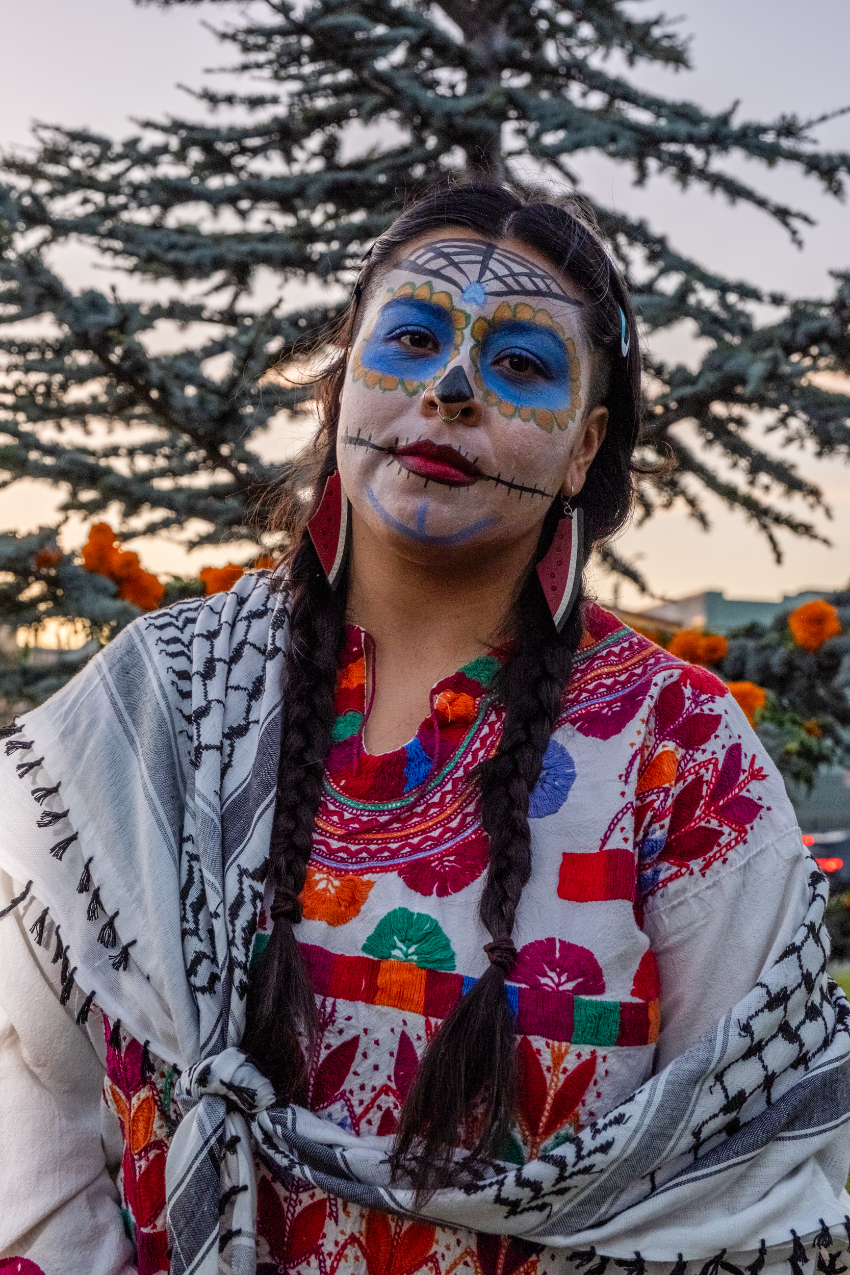 A person with vibrant Day of the Dead face paint and braided hair is wearing a colorful embroidered dress and shawl, standing in front of a tree with marigolds.