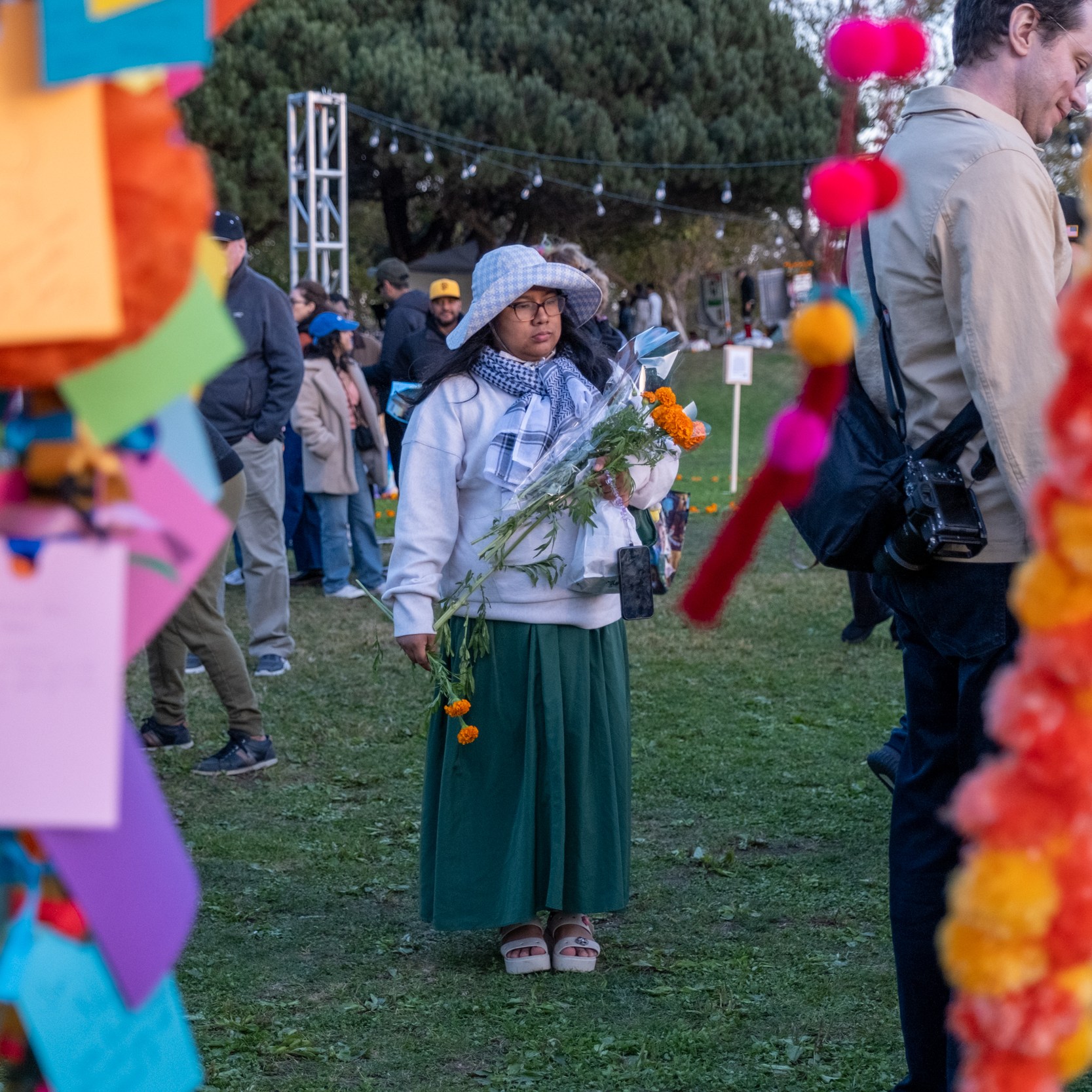 A person in a hat and scarf holds flowers at an outdoor event, surrounded by colorful notes and garlands. People stand in the background on a grassy area.