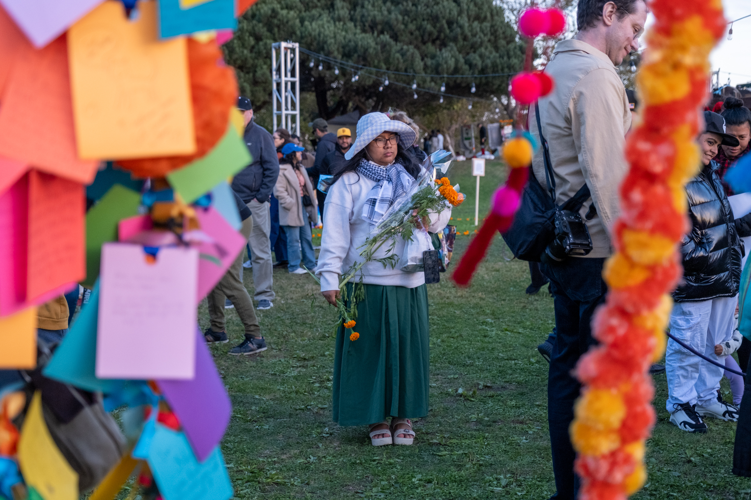 A person in a hat and scarf holds flowers at an outdoor event, surrounded by colorful notes and garlands. People stand in the background on a grassy area.