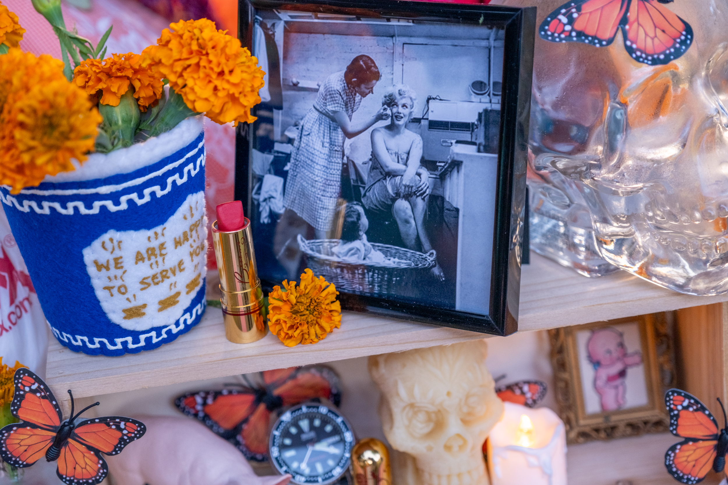 The image features a vintage black-and-white photo, marigold flowers, a lipstick, and a glass skull. Butterflies and a framed picture add decorative touches.