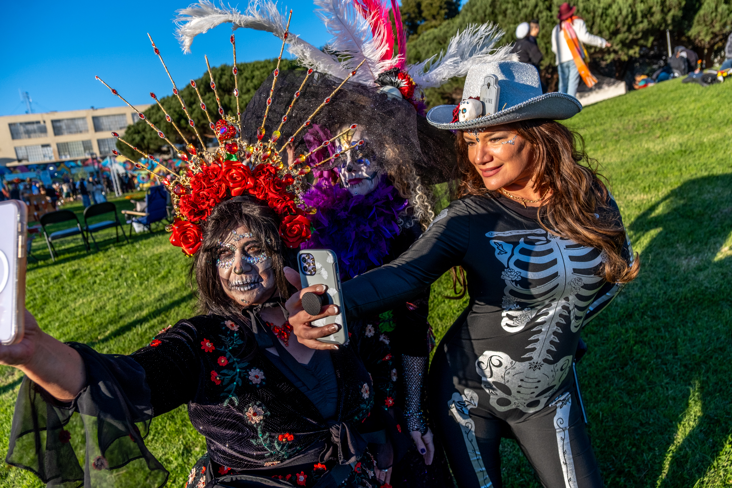 Three people in vibrant costumes pose for a selfie outdoors. One wears a red floral headdress, another has a purple feather boa, and the third dons a skeleton outfit.