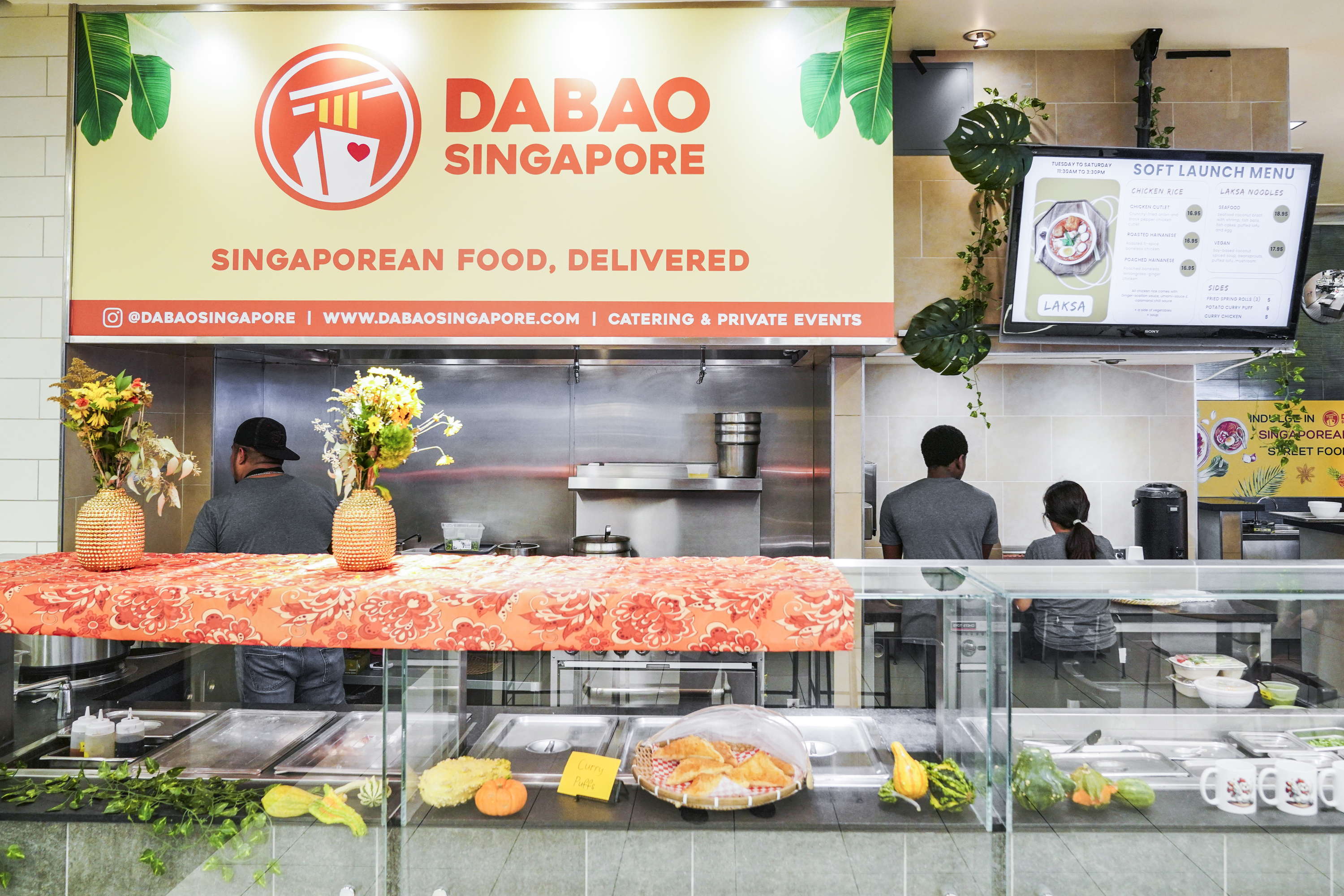 A vibrant food stall, &quot;Dabao Singapore,&quot; displays a menu and decor. Two staff work behind a counter adorned with flowers and food items, like curry puffs and produce.