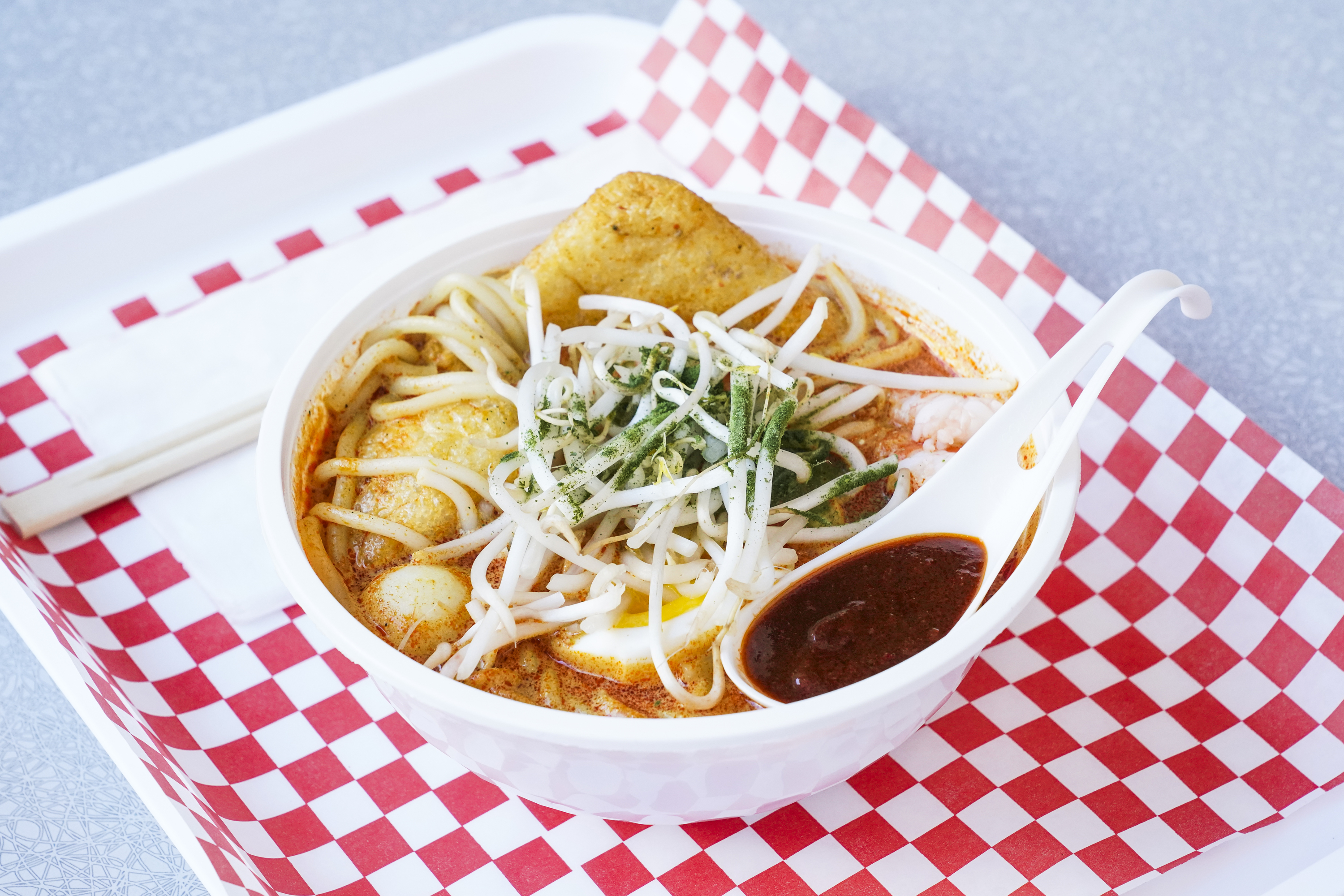 A bowl of noodle soup topped with bean sprouts, herbs, and a slice of fried tofu. A spoon holds a dark sauce, resting on a red-checkered tray with chopsticks.