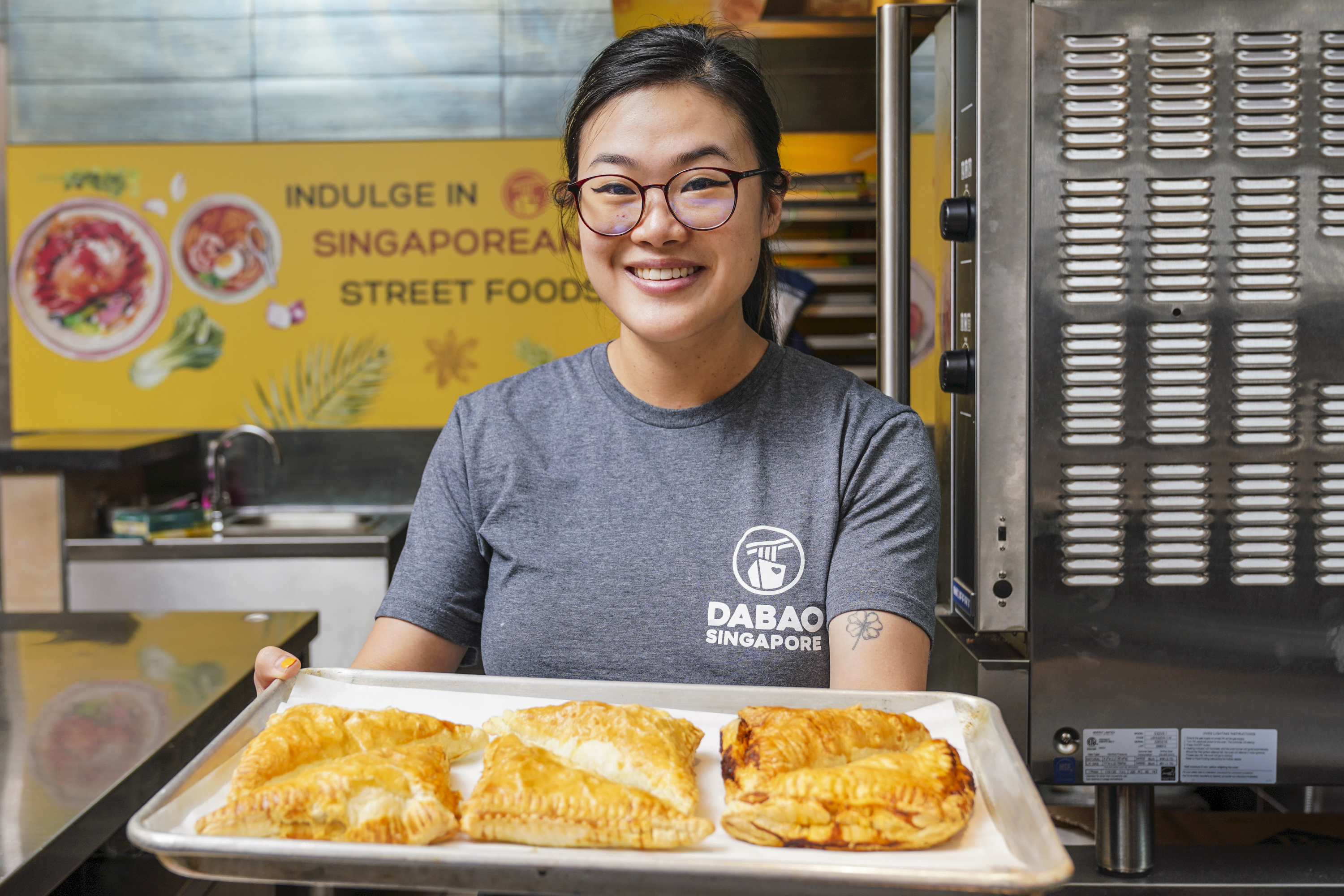 A smiling person in glasses holds a tray with baked pastries. Behind them is a yellow sign that reads "Indulge in Singaporean Street Food."