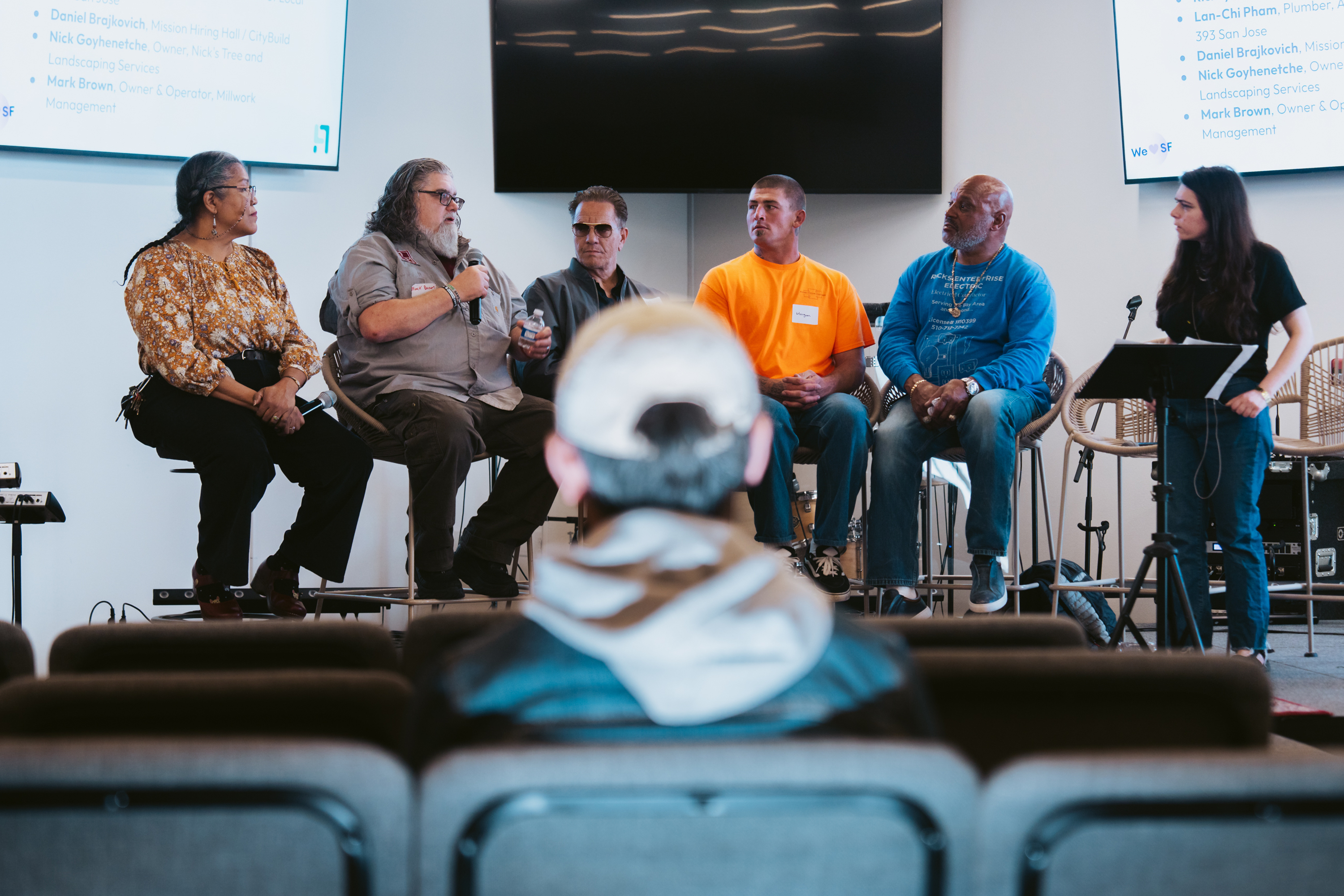 A panel of six people sits on stage in a discussion setting. One person holds a microphone, while an audience member is visible in the foreground.