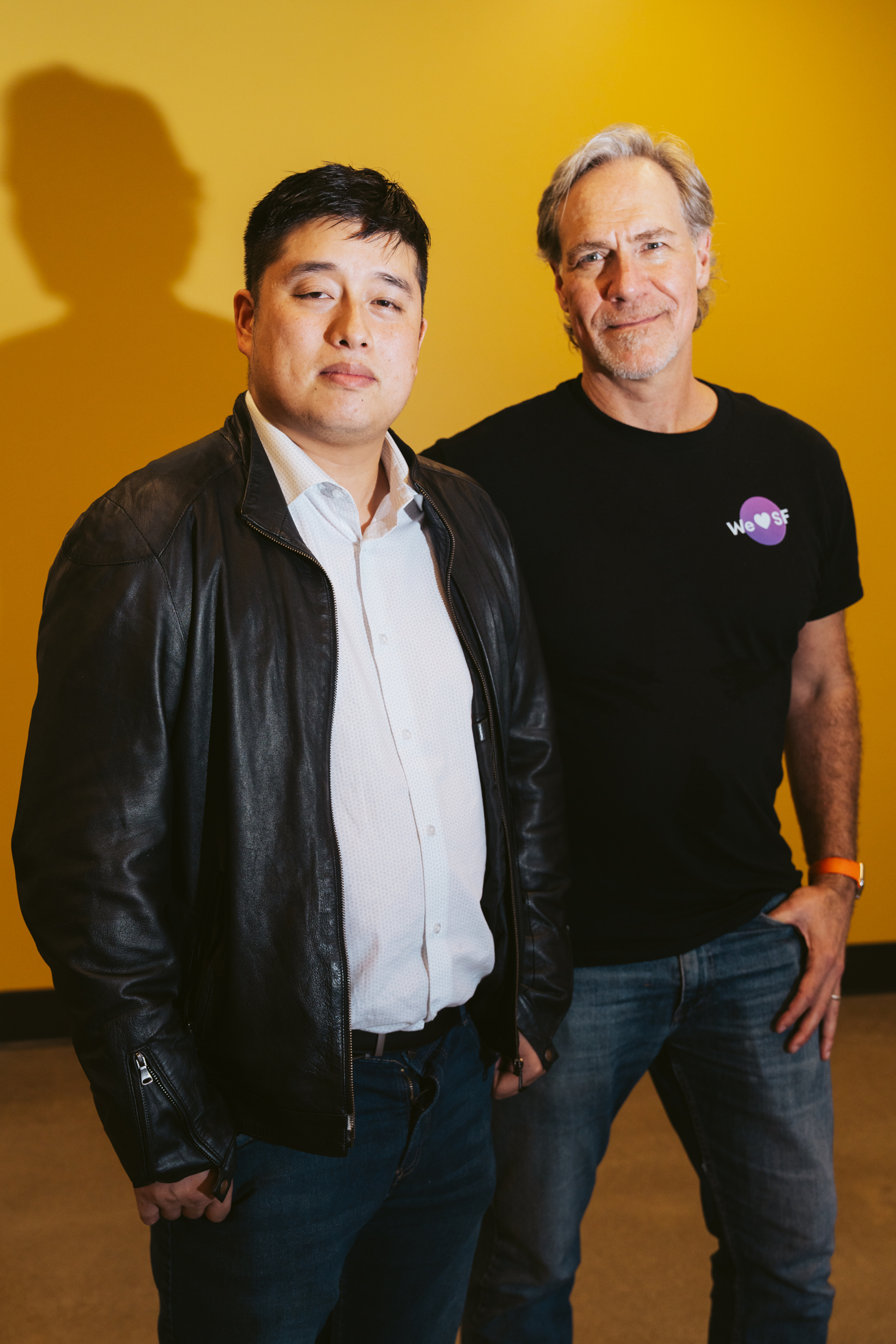 Two men stand side by side against a yellow background. One wears a leather jacket over a shirt, and the other wears a &quot;We ♥ SF&quot; T-shirt and jeans.