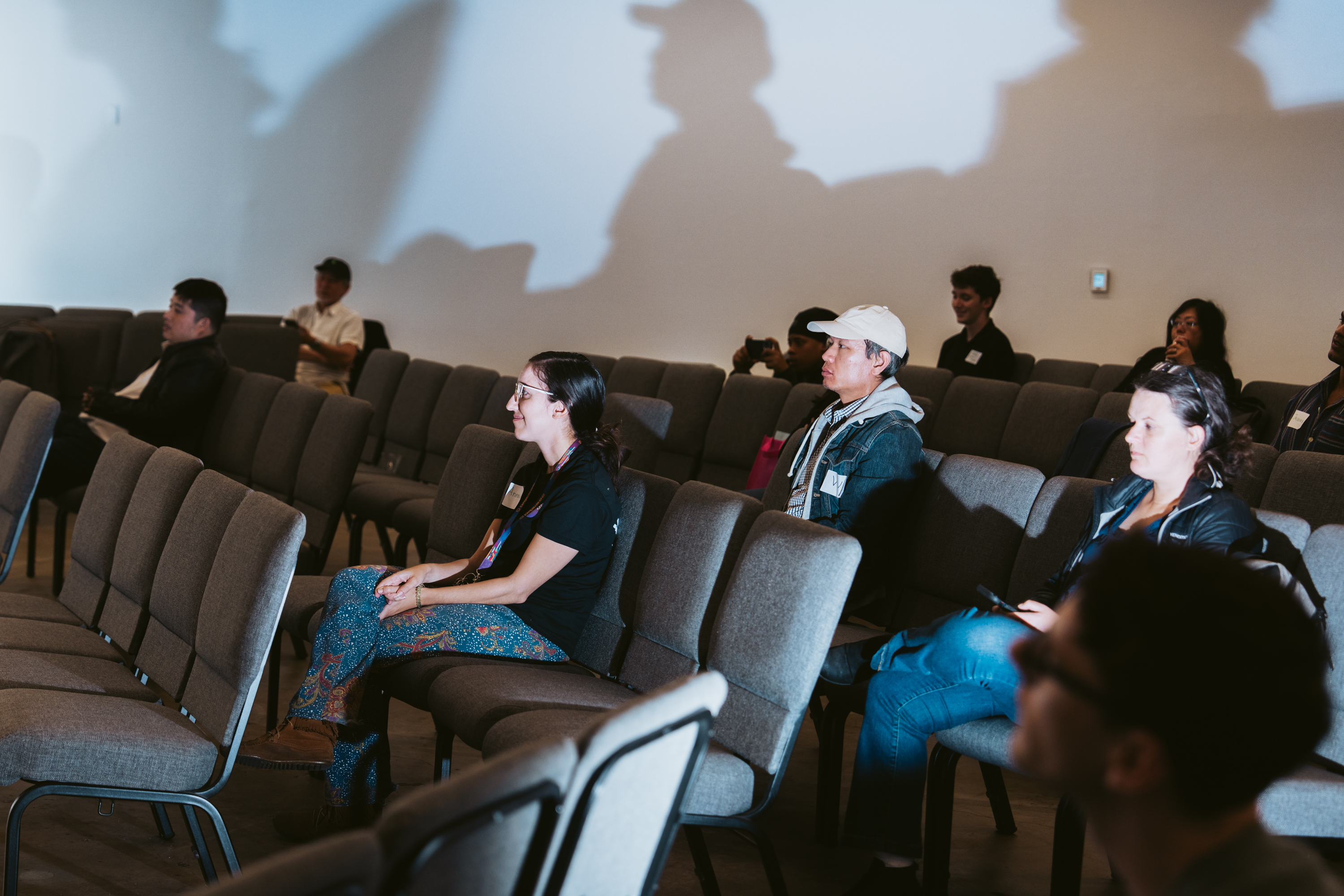 A group of people sit scattered in a dimly lit room with rows of empty chairs. Shadows cast on the wall above them create an intriguing visual effect.