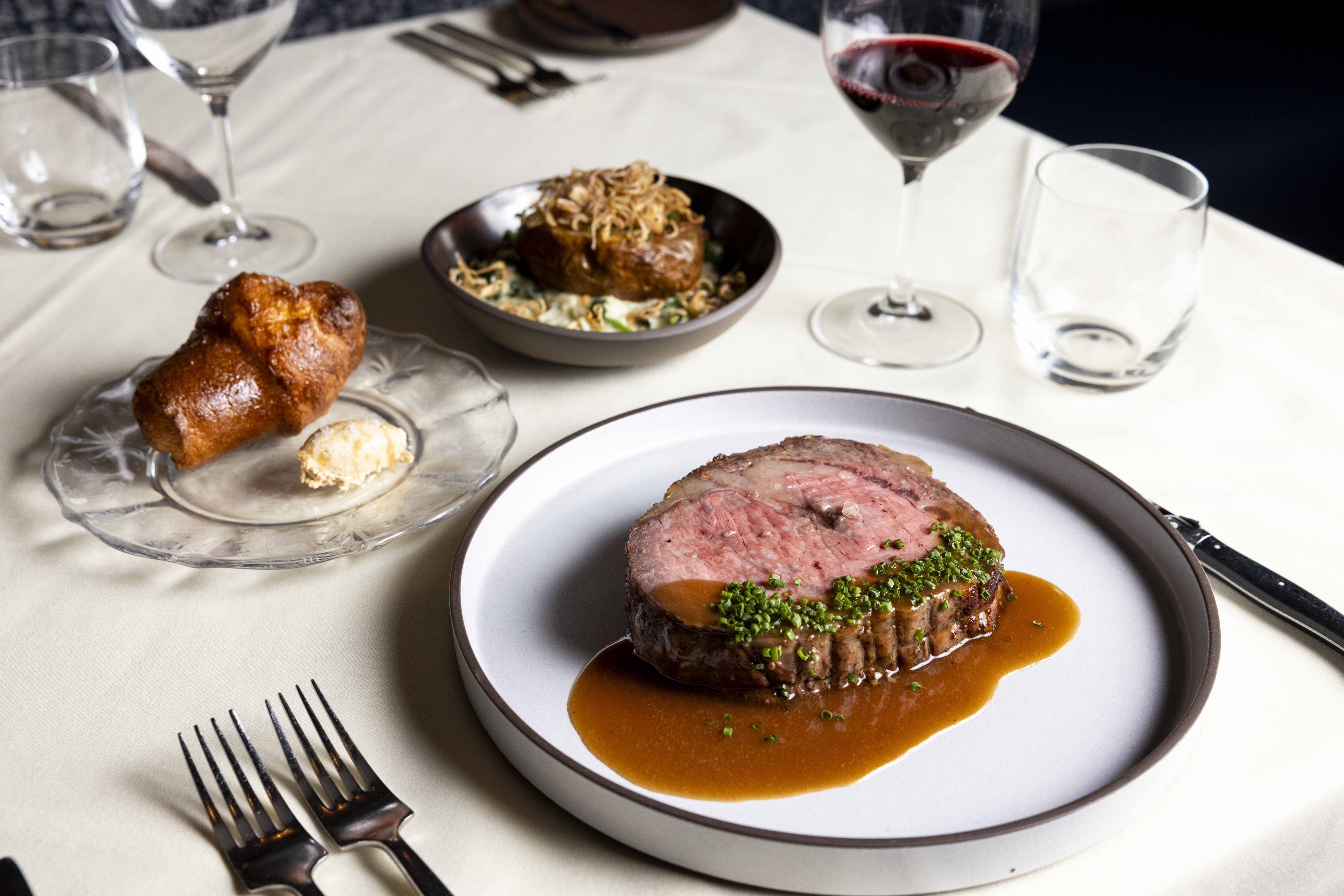 A plate with a slice of roast beef, chives, and sauce; a popover with butter; a bowl with a stuffed potato; and a glass of red wine on a white tablecloth.