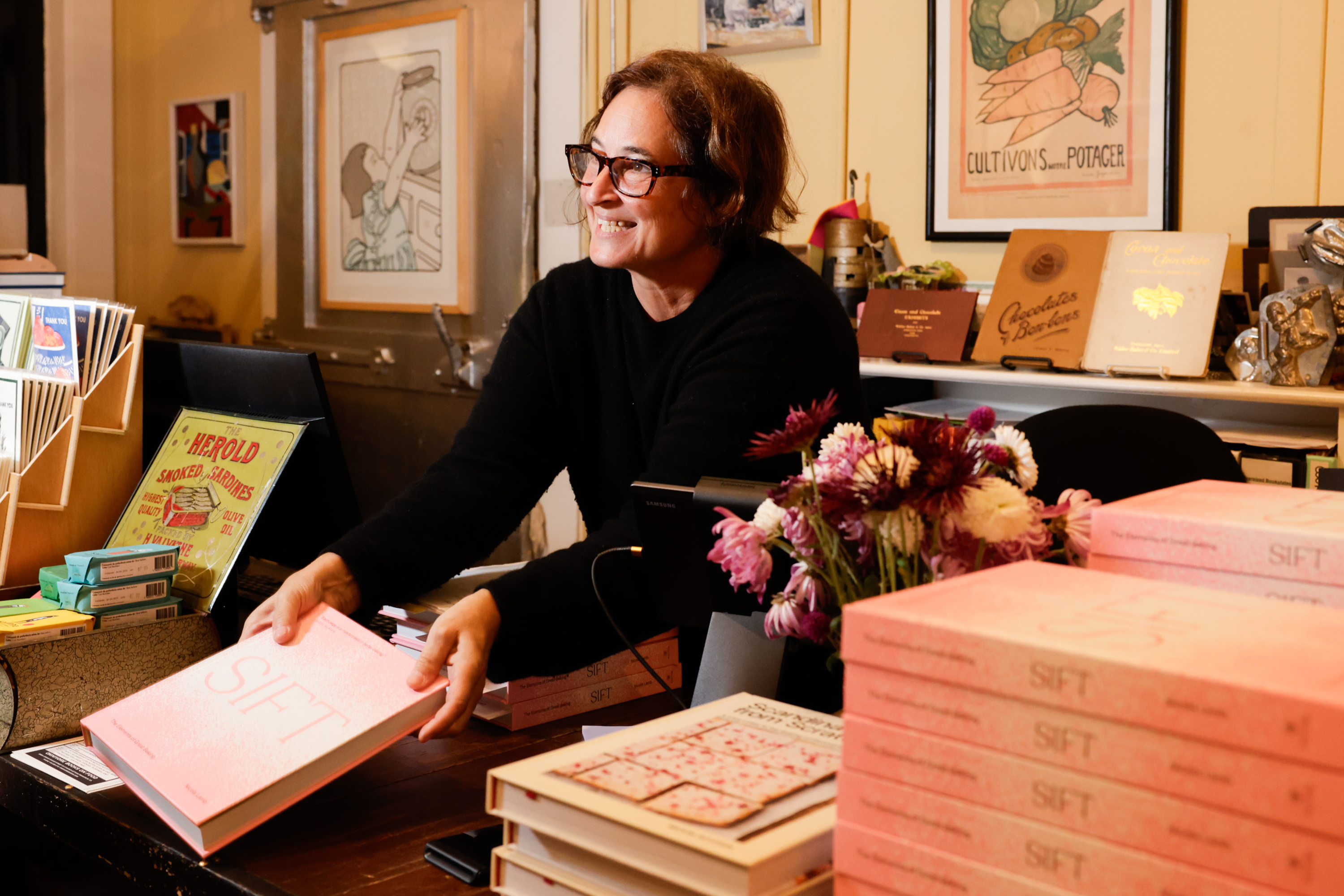 A woman with glasses smiles behind a counter filled with pink books titled &quot;SIFT.&quot; There are flowers nearby and various posters and items in the background.