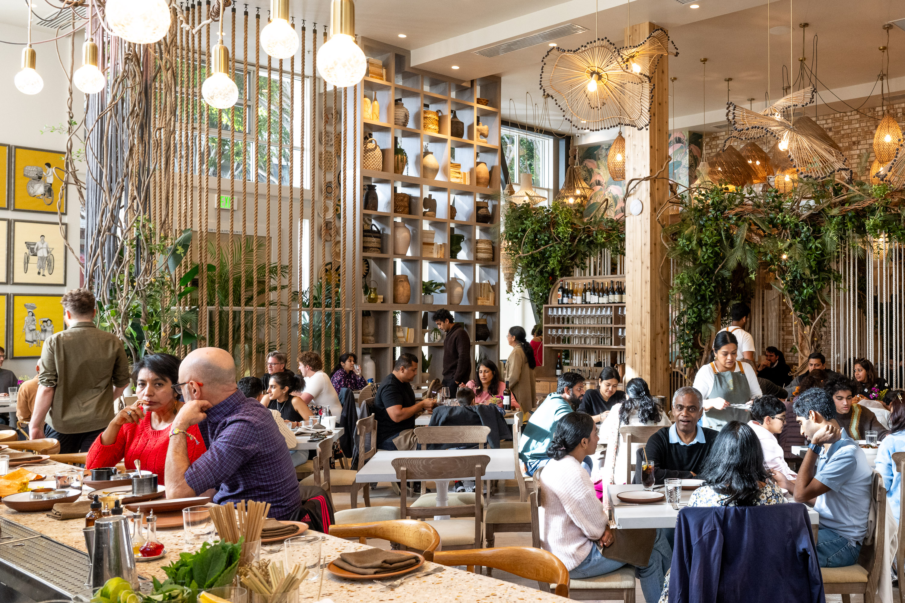 A bustling restaurant with people dining at tables, surrounded by plants and shelves of pottery. Warm lighting creates a cozy atmosphere, and servers attend to guests.