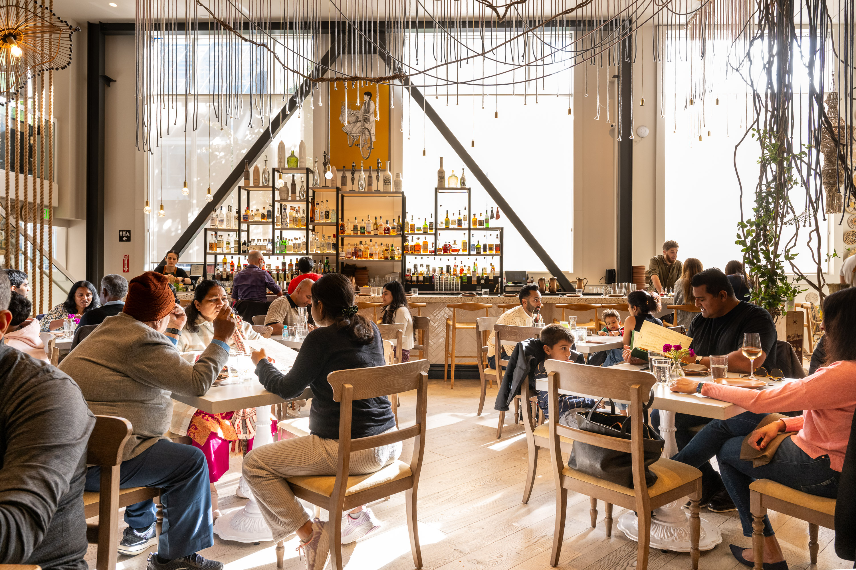 The image shows a lively restaurant filled with people seated at tables, engaged in conversation. The backdrop features shelves with various bottles and elegant decor.