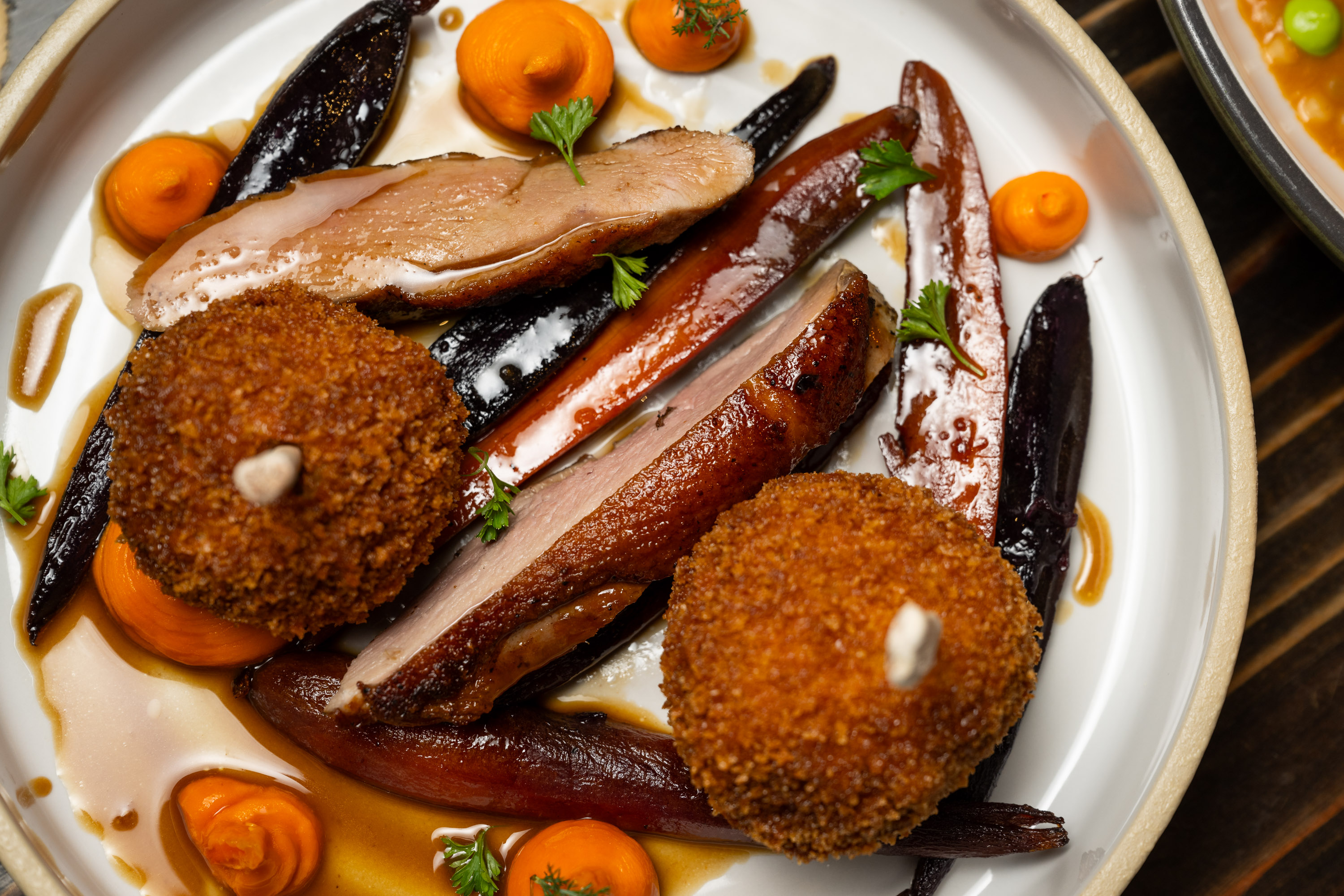 The image shows a gourmet dish with sliced duck breast, crispy golden croquettes, colorful carrots, carrot puree, and fresh herbs on a white plate.