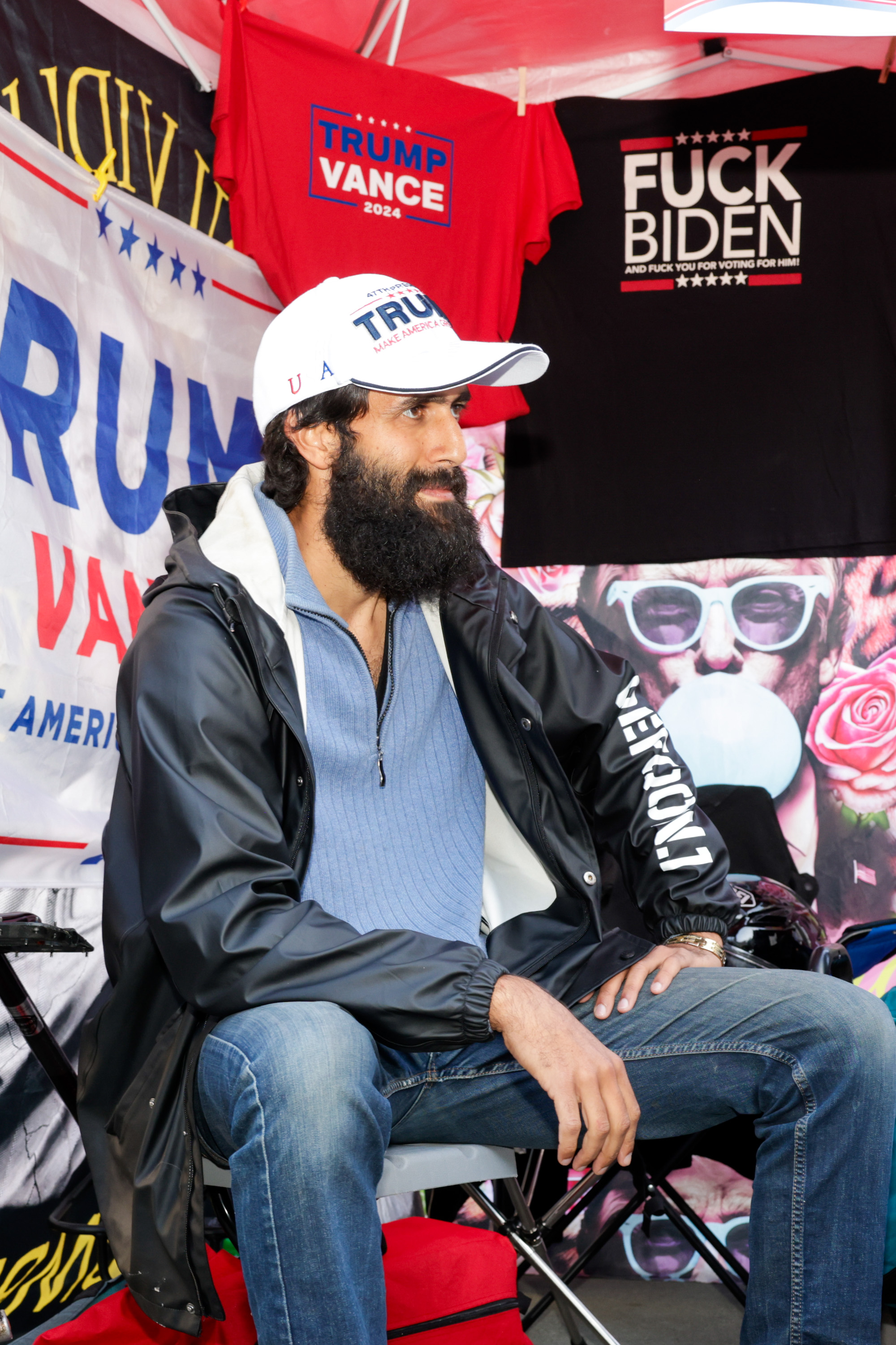 A bearded man in a &quot;Trump&quot; hat sits in front of political merchandise, including shirts with slogans and a Trump Vance banner, under a red canopy.