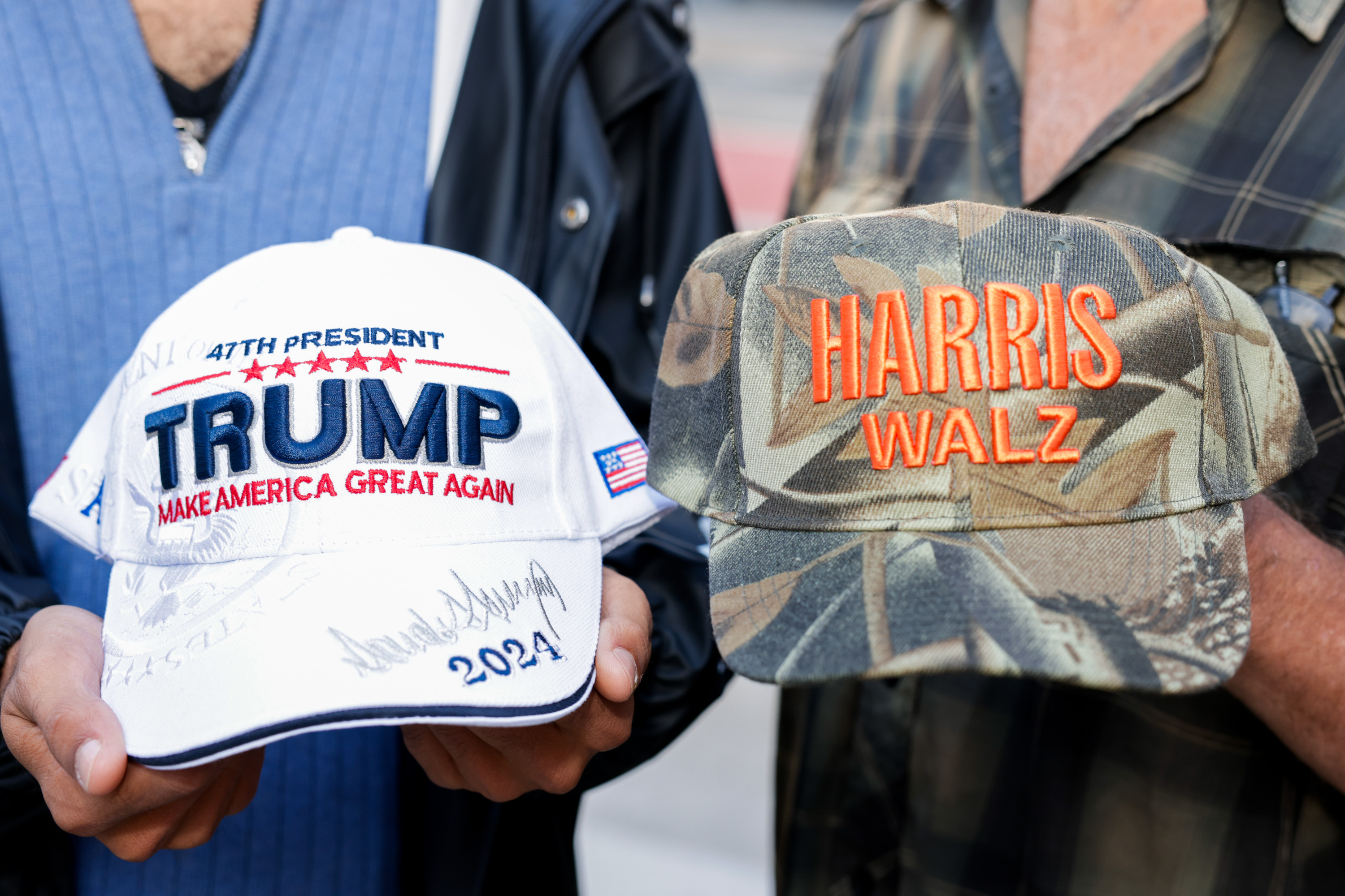 Two people hold caps: one white with &quot;TRUMP&quot; and &quot;2024&quot;, and one camo with &quot;HARRIS WALZ&quot; in orange.