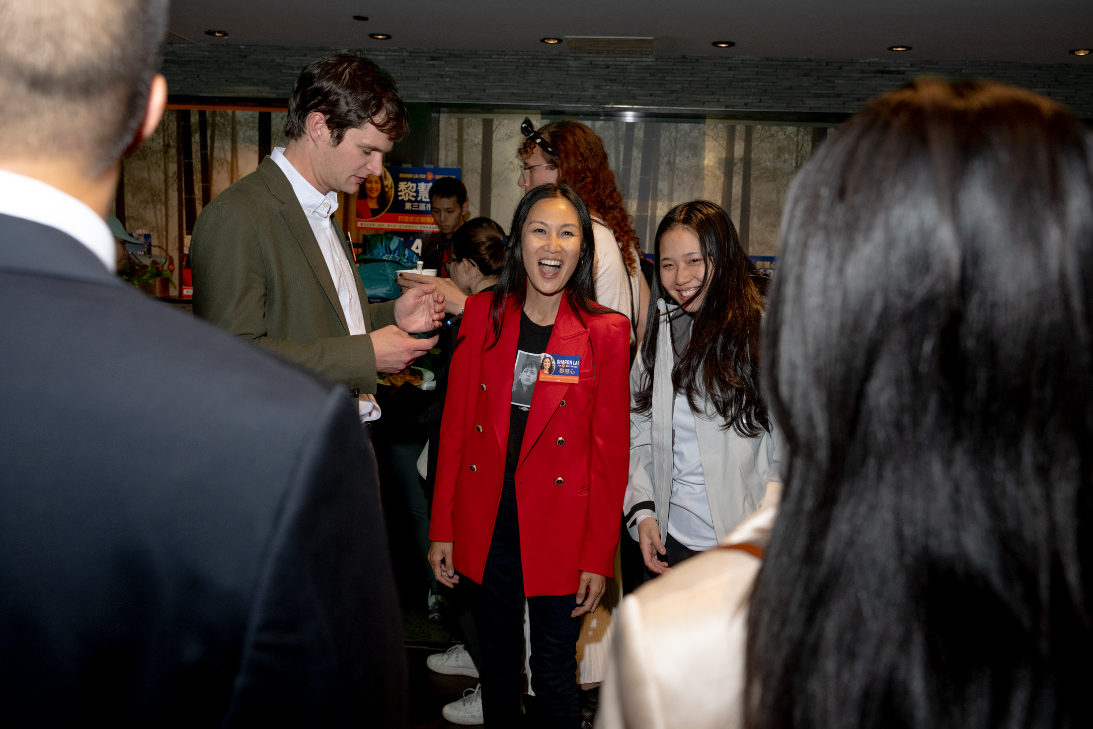 A group of people are gathered indoors, smiling and engaging. A woman in a red blazer stands prominently, while others converse and check phones around her.