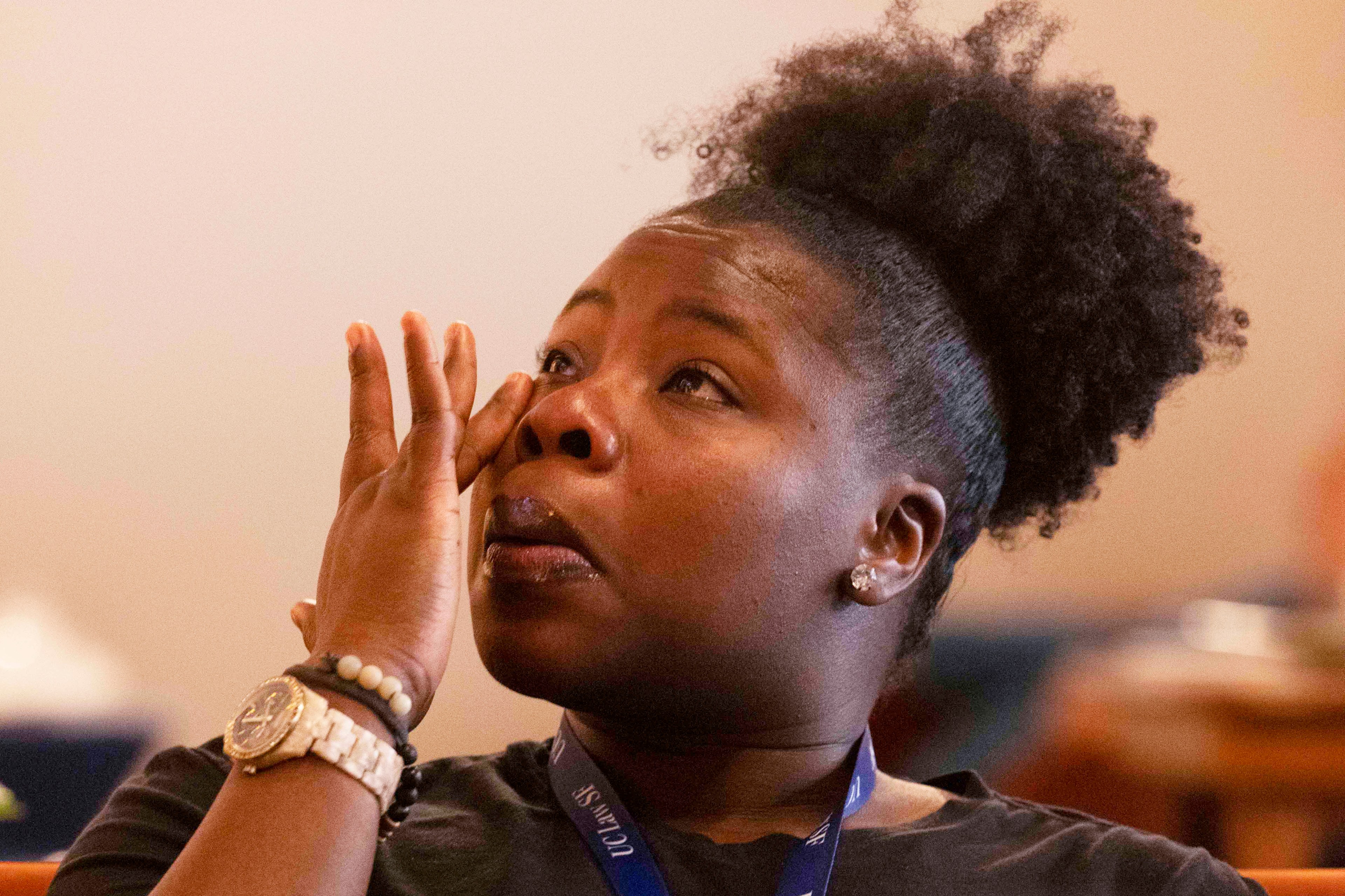 A woman with a watch and bracelets wipes her eye. Her hair is pulled back into a puff, and she wears a lanyard around her neck, appearing emotional.