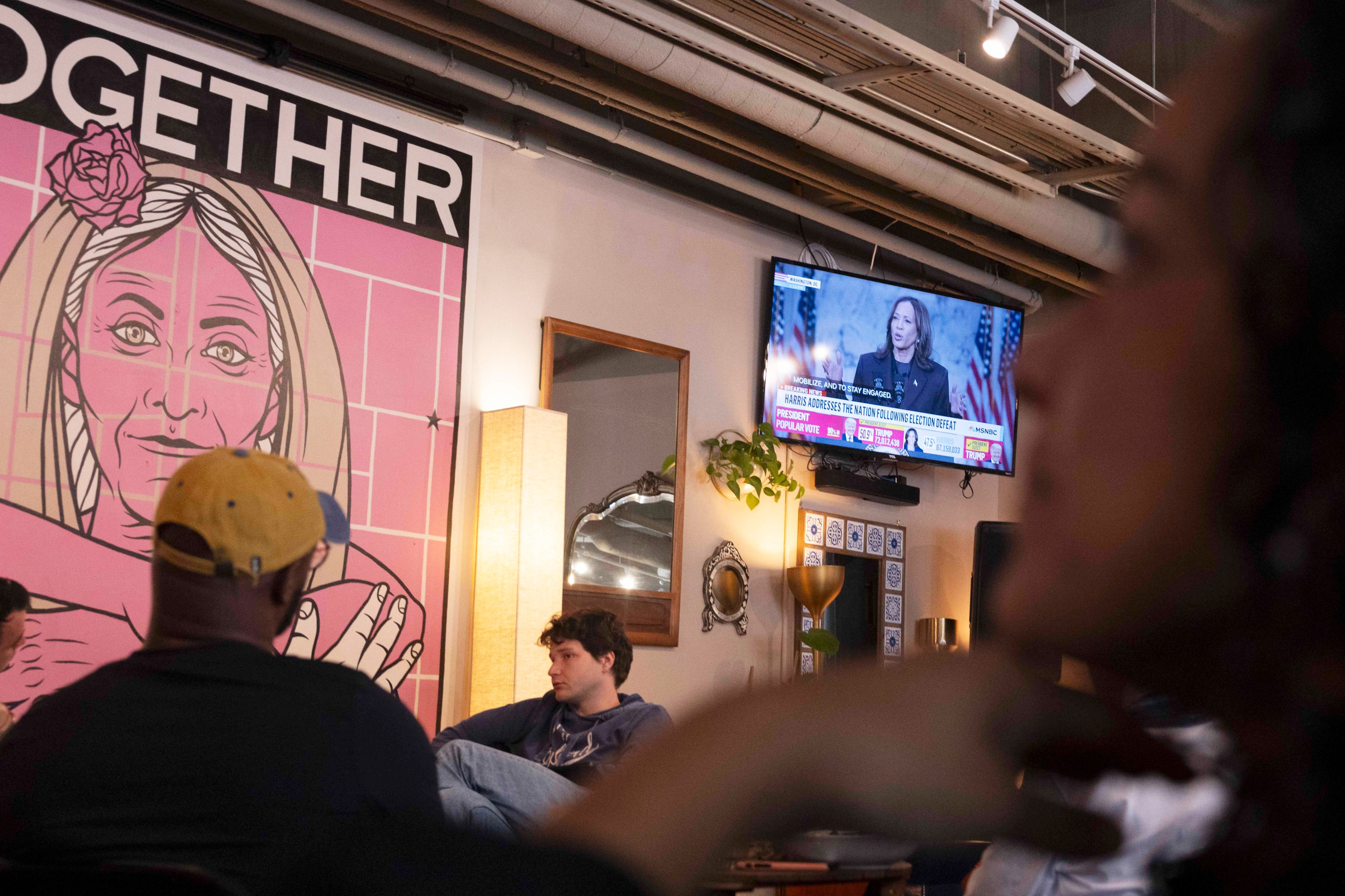 A casual setting with people seated, featuring a large pink mural of a person alongside a TV showing a news broadcast. The space is warmly lit.