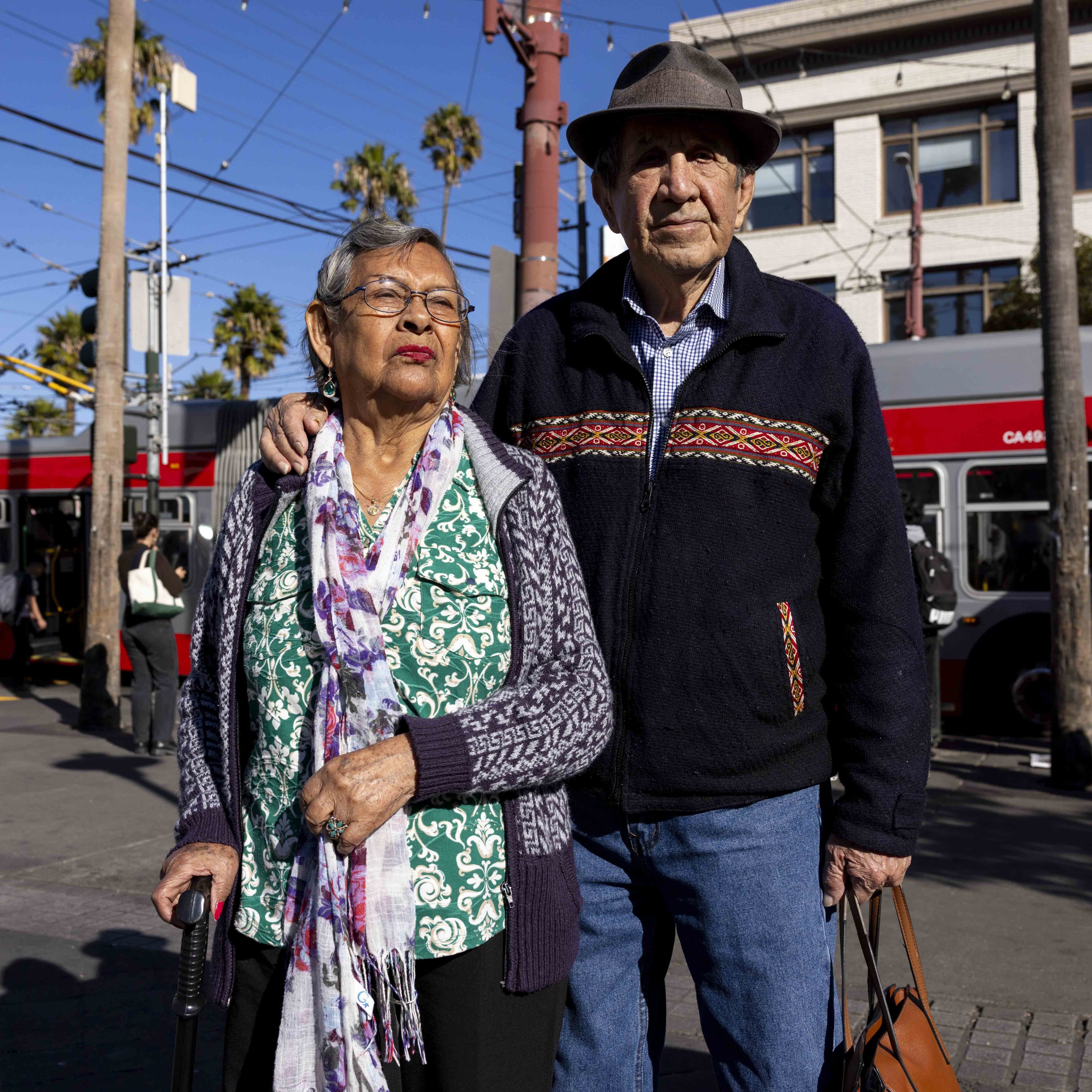 An elderly couple stands together in a sunny urban setting. The woman holds a cane, and the man wears a hat. A red bus and pedestrians are in the background.