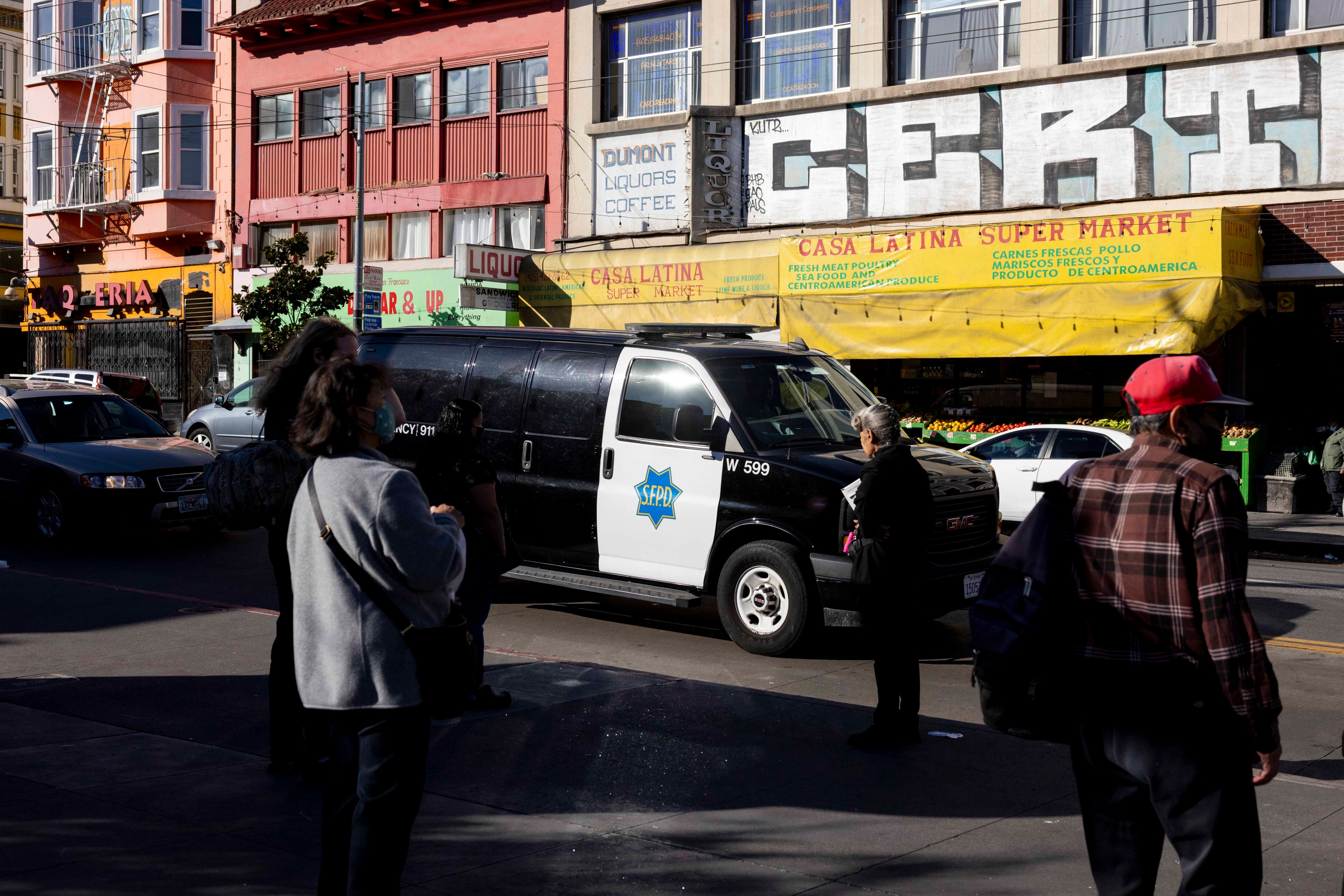 The image shows a street scene with a black and white police van marked "SFPD" parked near storefronts, including a market and liquor store, with people walking by.