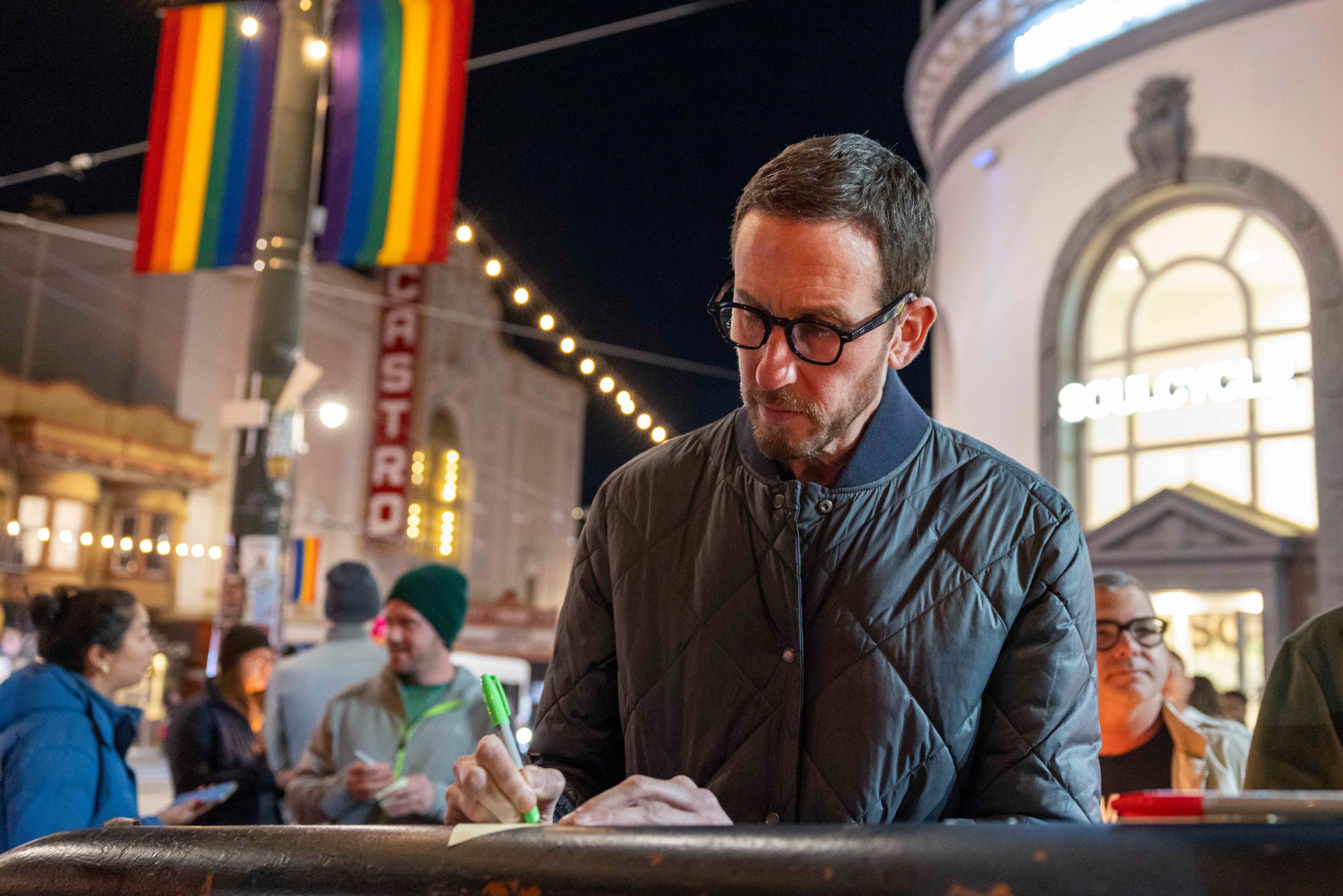 A man in glasses and a quilted jacket writes with a green pen at a nighttime street event. Rainbow flags and festive lights adorn the background.