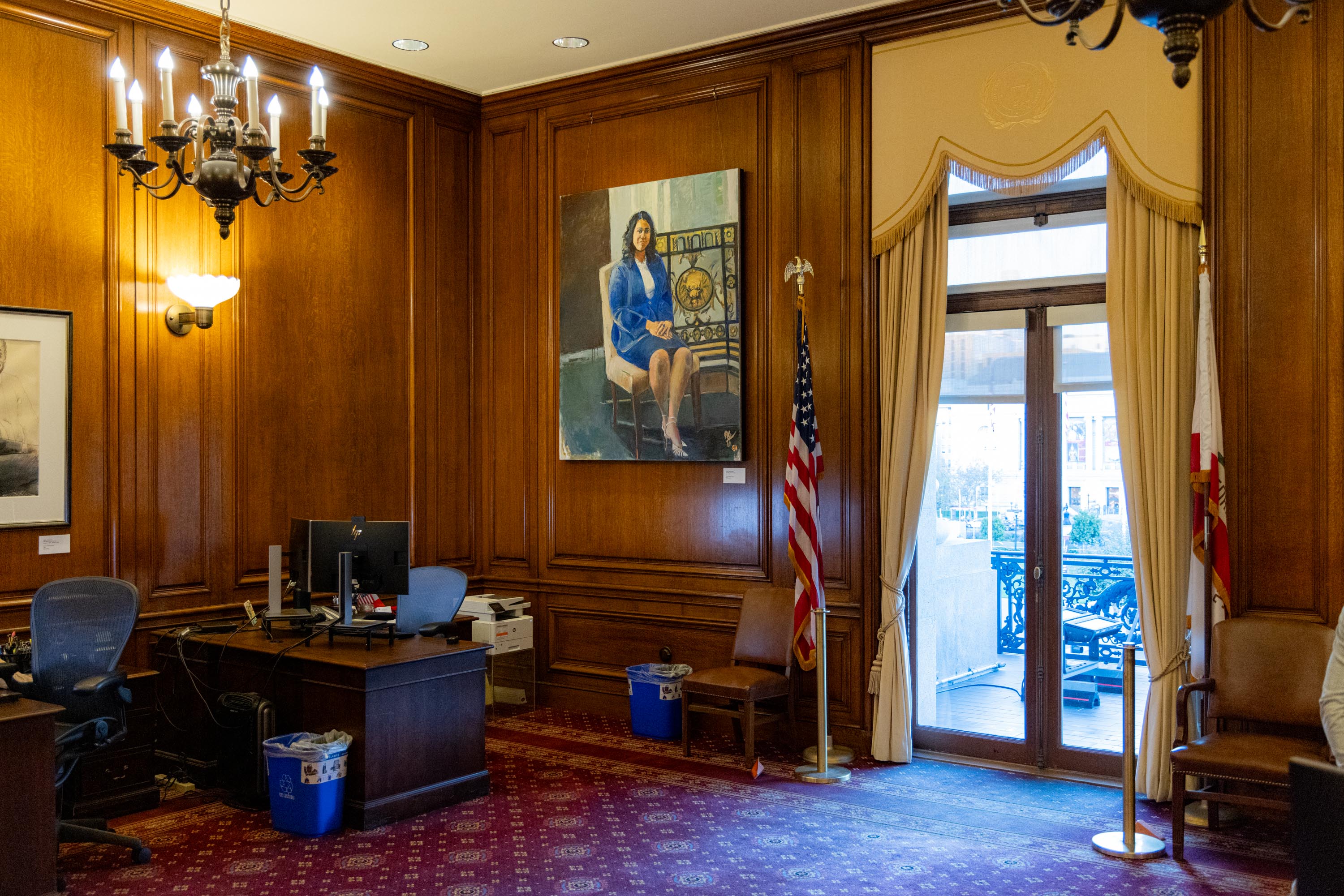 The room features wood-paneled walls, a desk with computers, a large portrait, an American flag, and glass doors leading to a balcony with city views.