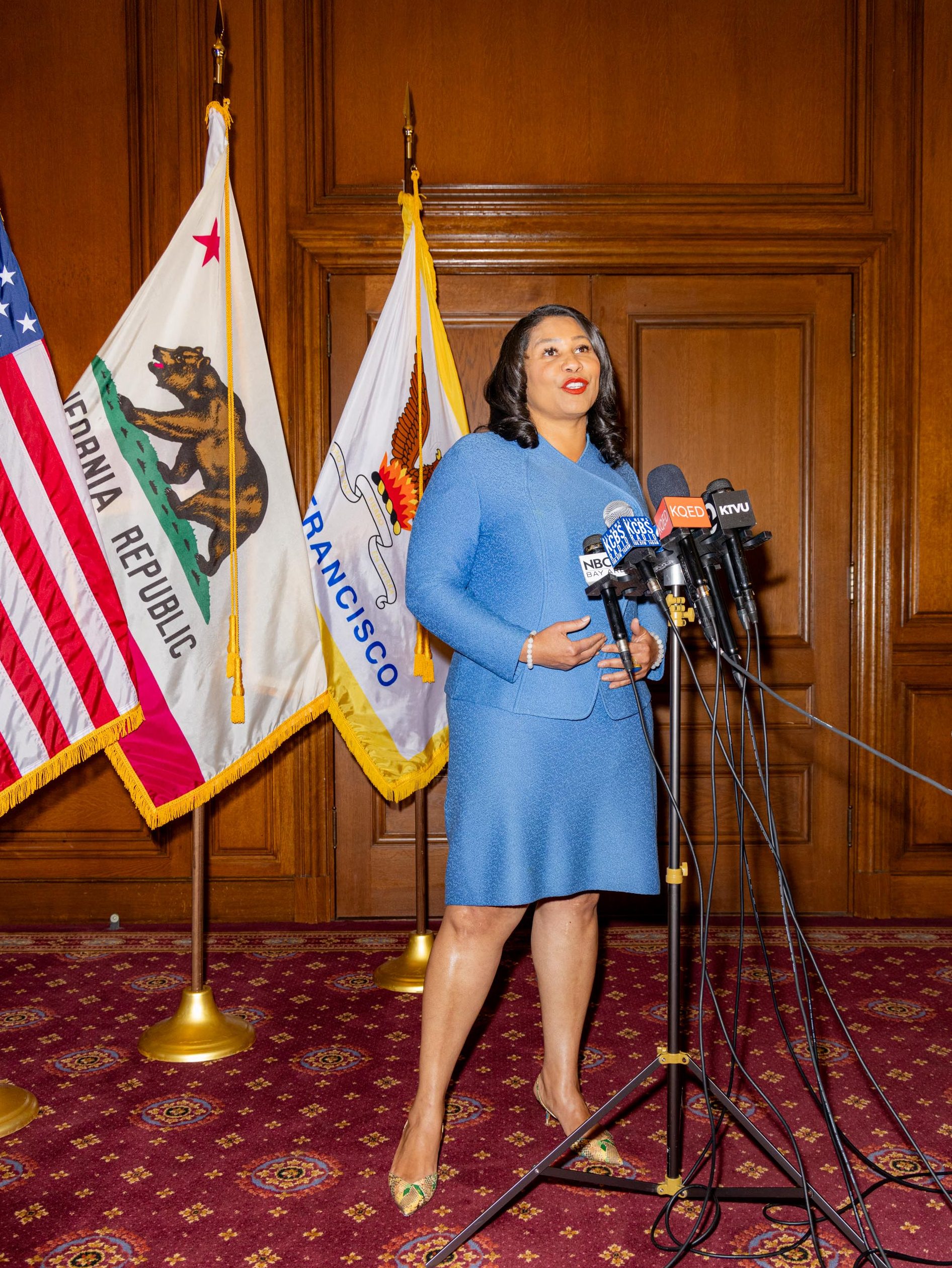 A woman in a blue suit stands at a podium with microphones. Flags with California and San Francisco symbols are behind her in a wood-paneled room.