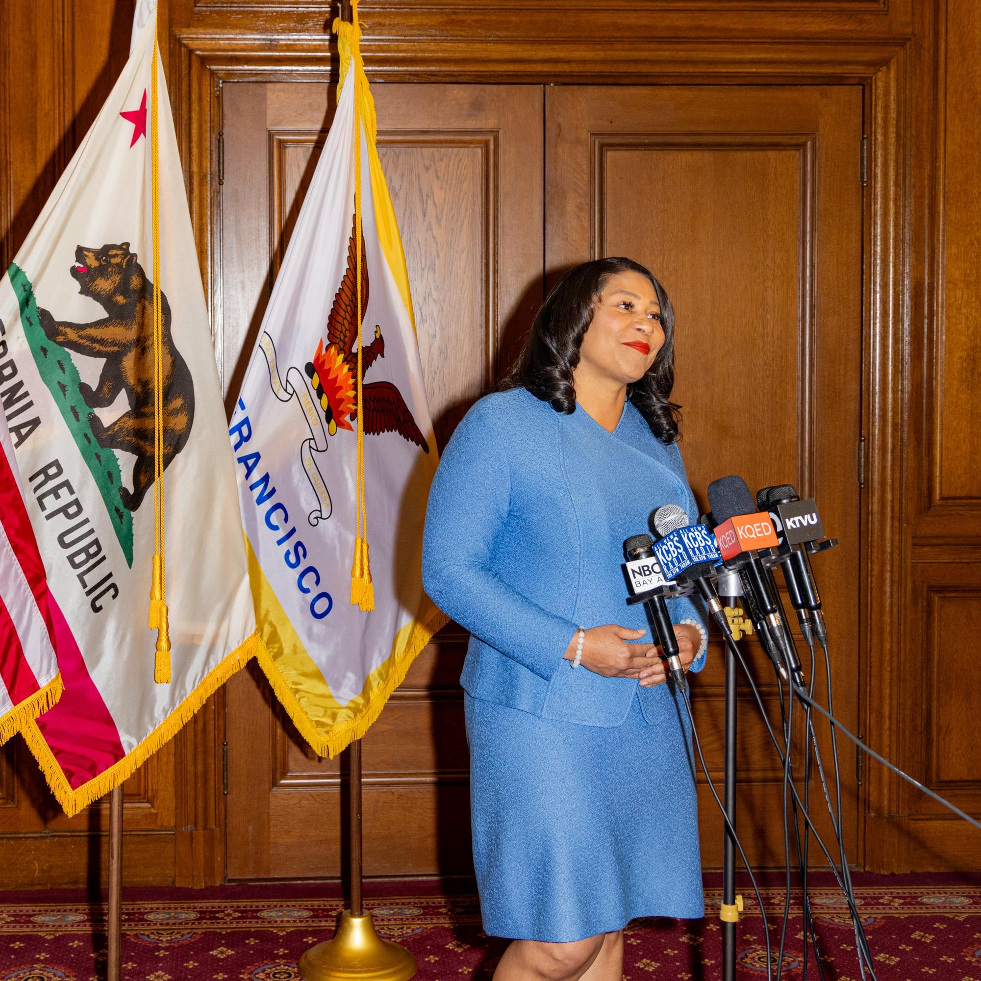 A person in a blue suit stands at a podium with multiple microphones, flanked by the U.S., California, and San Francisco flags inside a wood-paneled room.