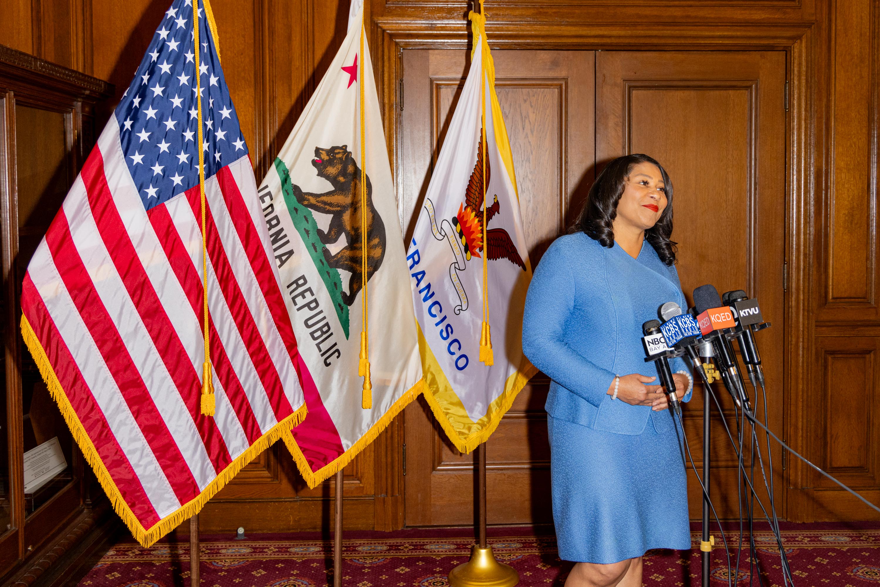 A person in a blue suit stands at a podium with multiple microphones, flanked by the U.S., California, and San Francisco flags inside a wood-paneled room.