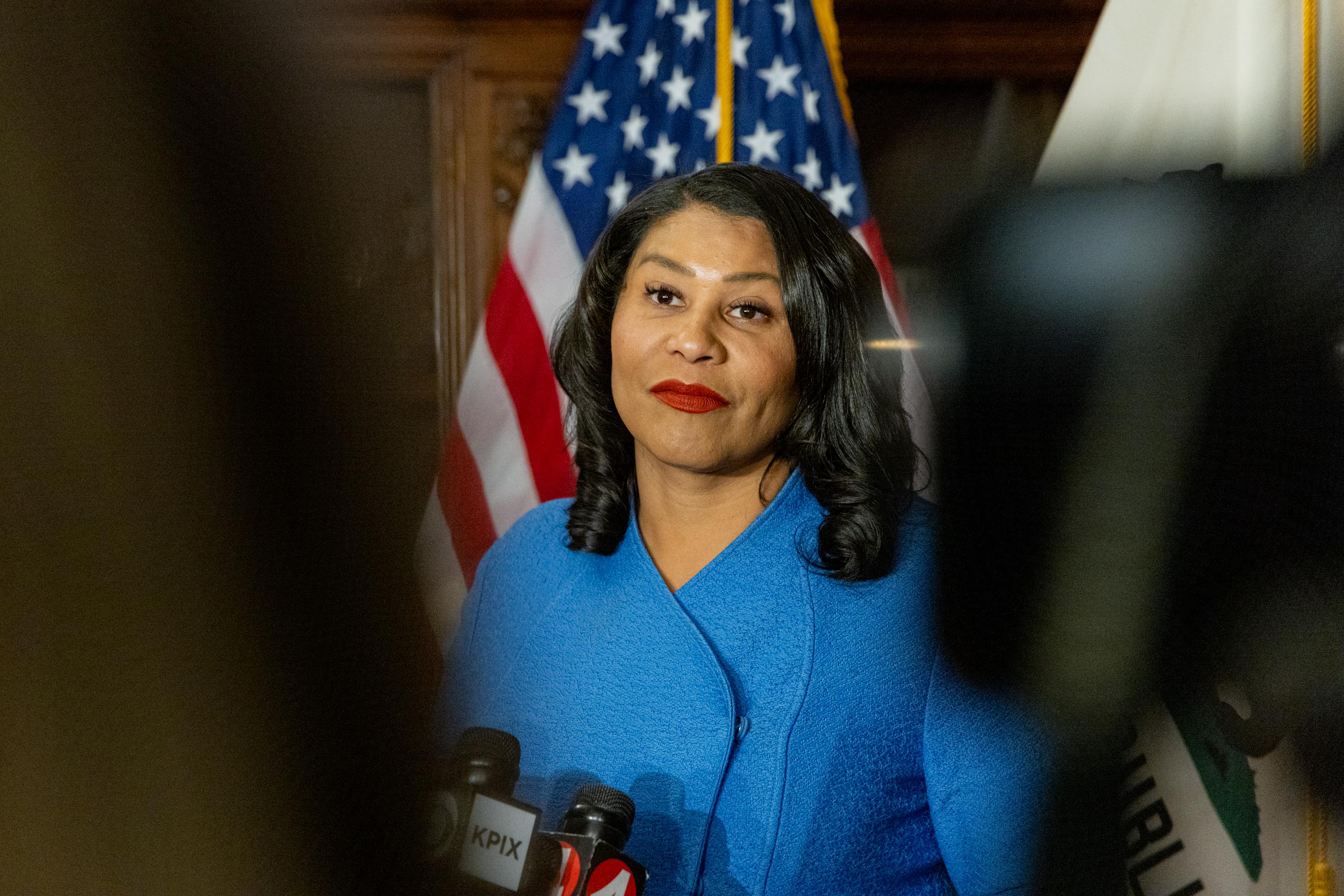 Mayor Breed seen in a blue jacket in a photo from the midsection-up with an American flag behind her. She's in the mayor's office, which is wood paneled.