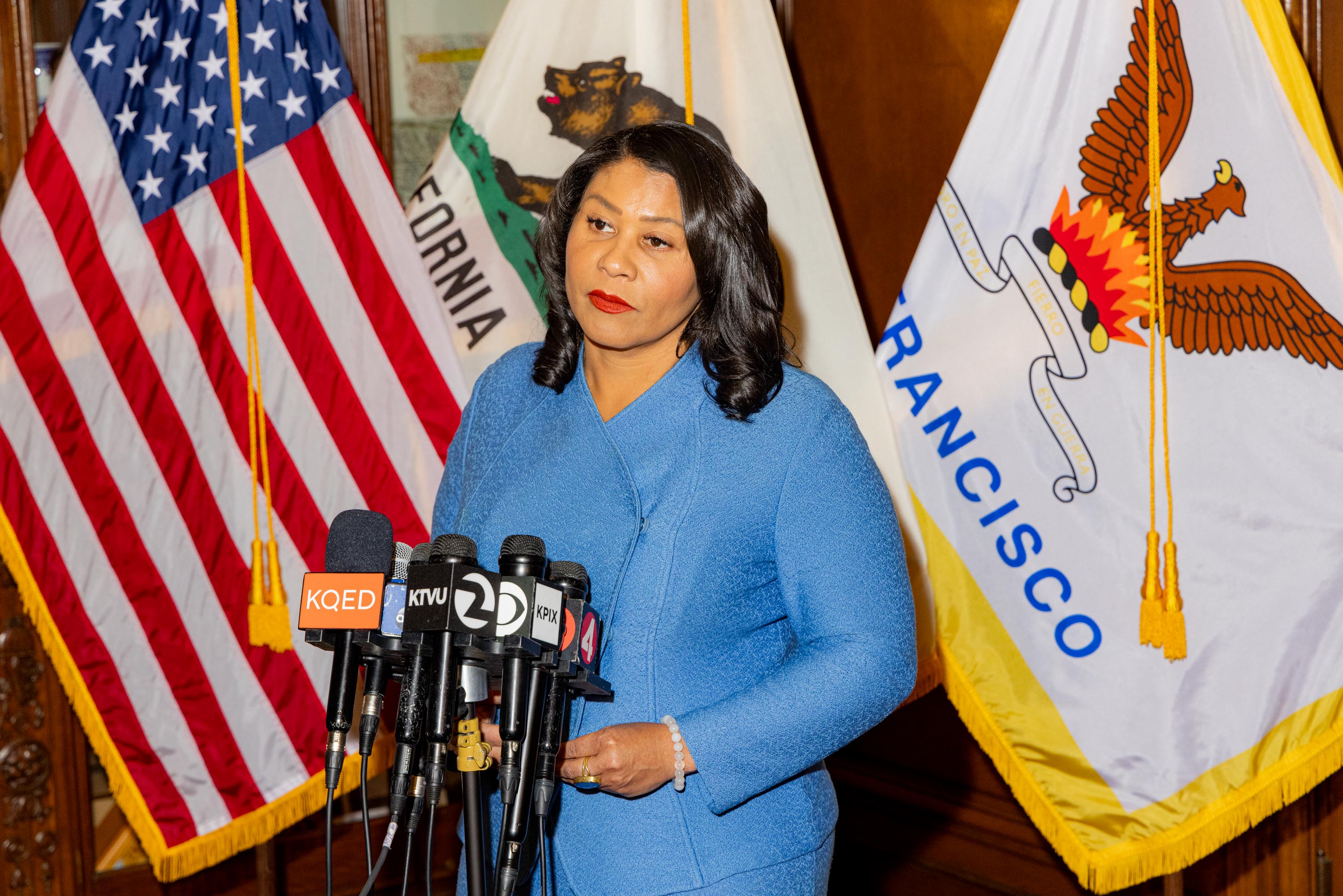 A woman in a blue jacket stands before a row of microphones, with American, California, and San Francisco flags behind her.