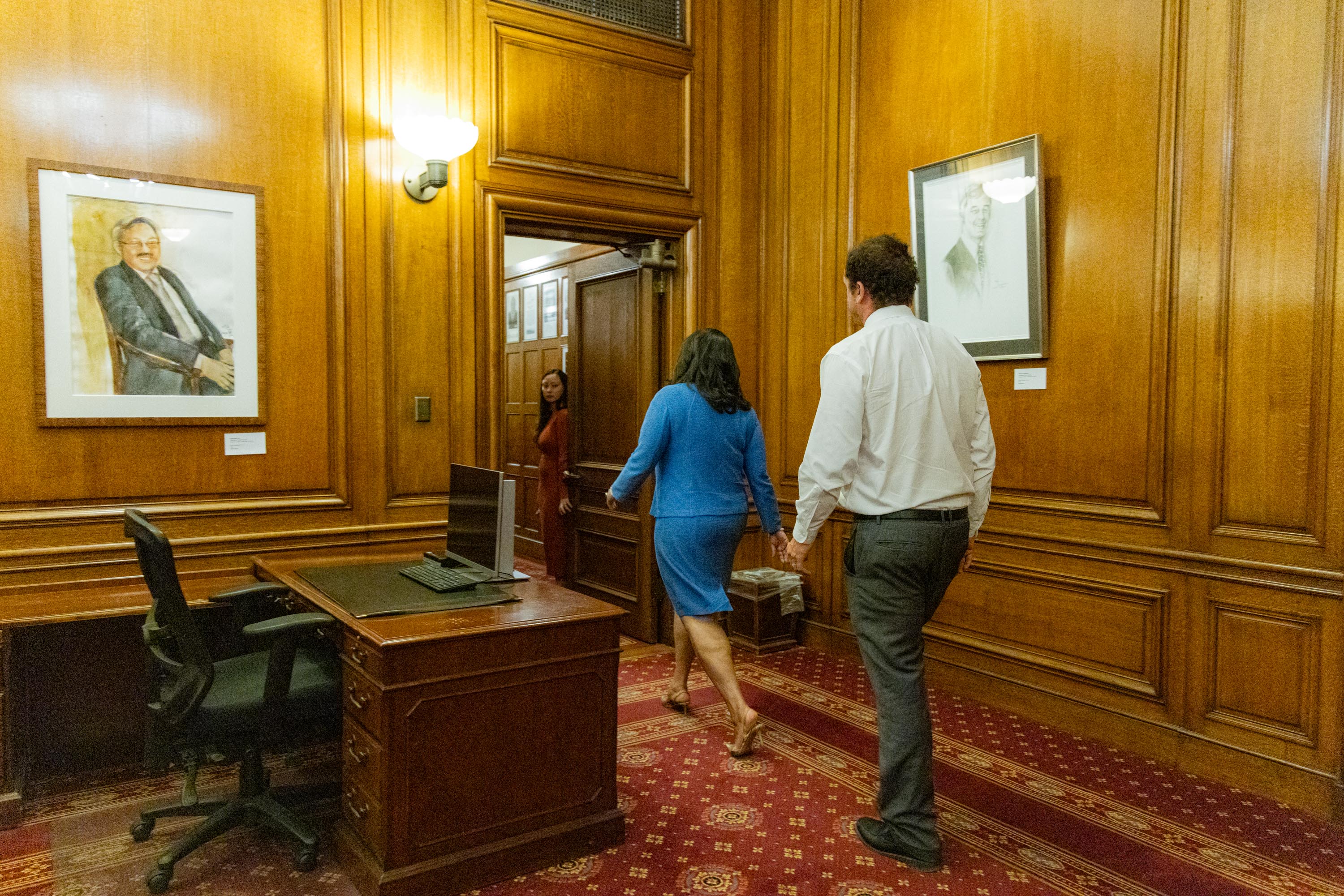 Three people walk through a wood-paneled office with a desk and chairs, adorned with framed portraits on the walls and a red carpet on the floor.