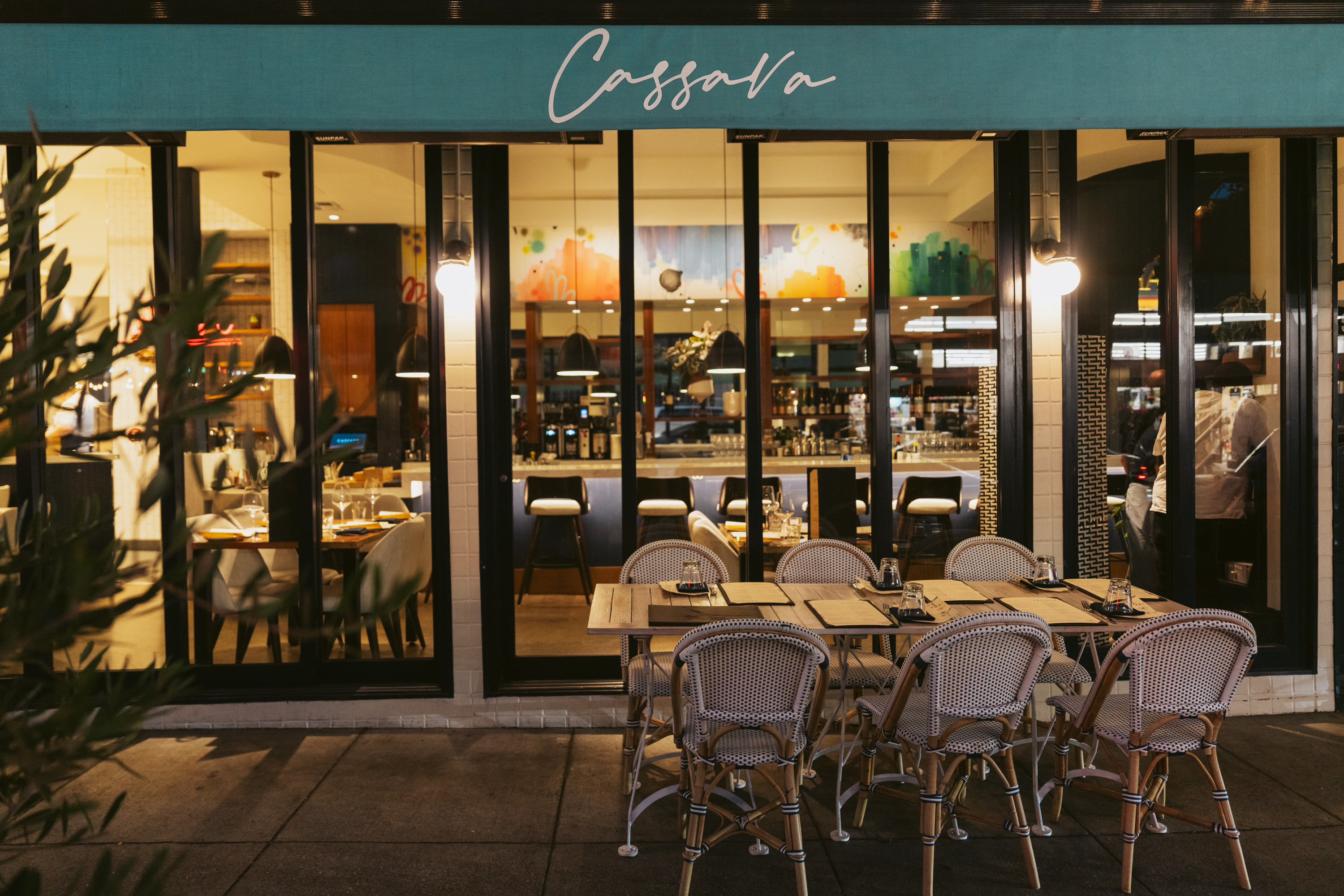 The image shows a restaurant with a teal awning labeled &quot;Cassava.&quot; It features elegant table settings outside and a warmly lit interior with a bar and colorful decor.