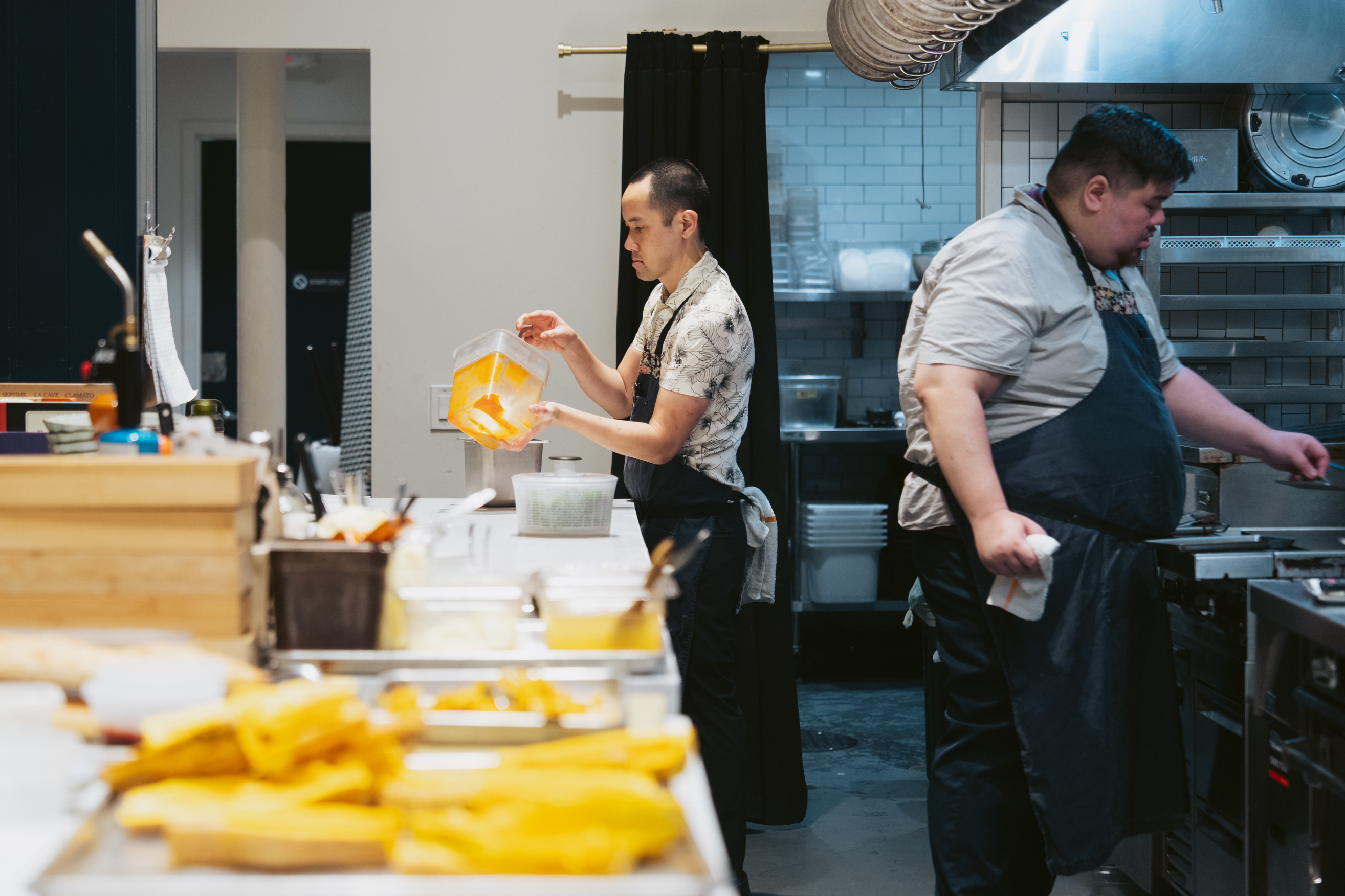 Two chefs are busy working in a kitchen. One is pouring ingredients from a container, while the other is focused on cooking at a stove. Various supplies are visible.