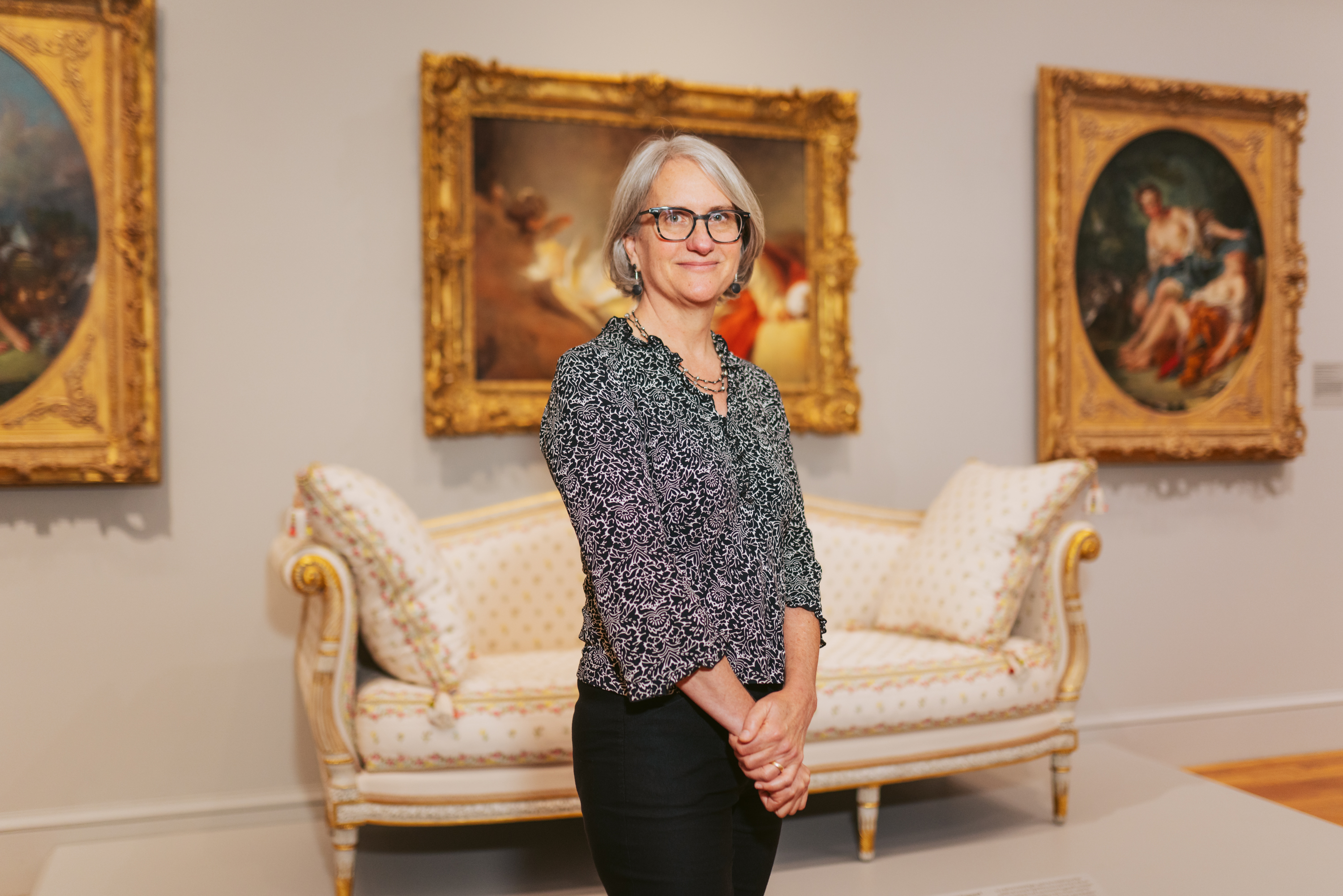 A person with gray hair and glasses stands in an art gallery, wearing a patterned shirt. Behind them is a vintage sofa and ornate framed paintings on the walls.