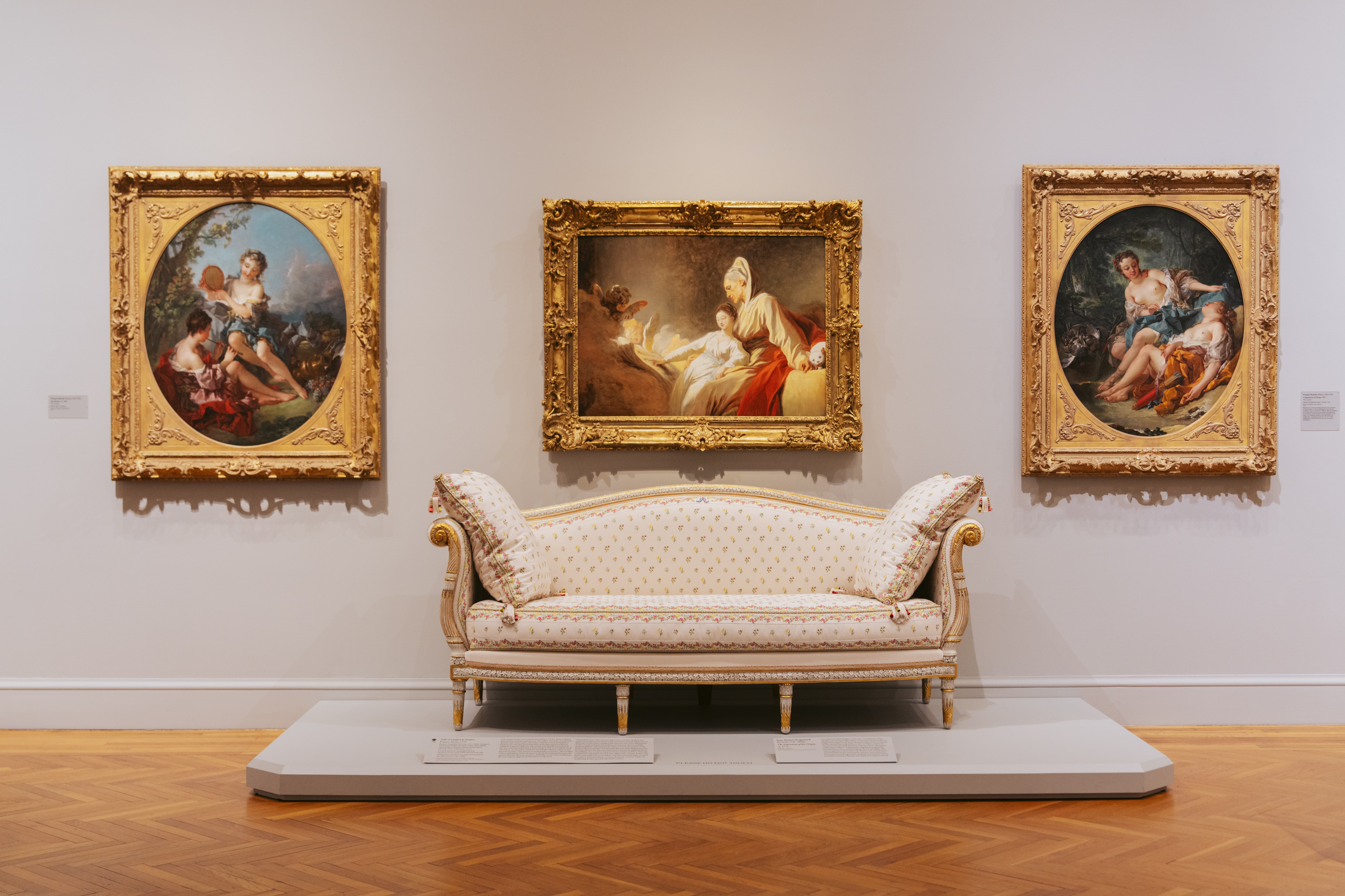 The image shows a museum display with an ornate sofa centered beneath three framed classical paintings on a white wall. The floor is wooden with a herringbone pattern.