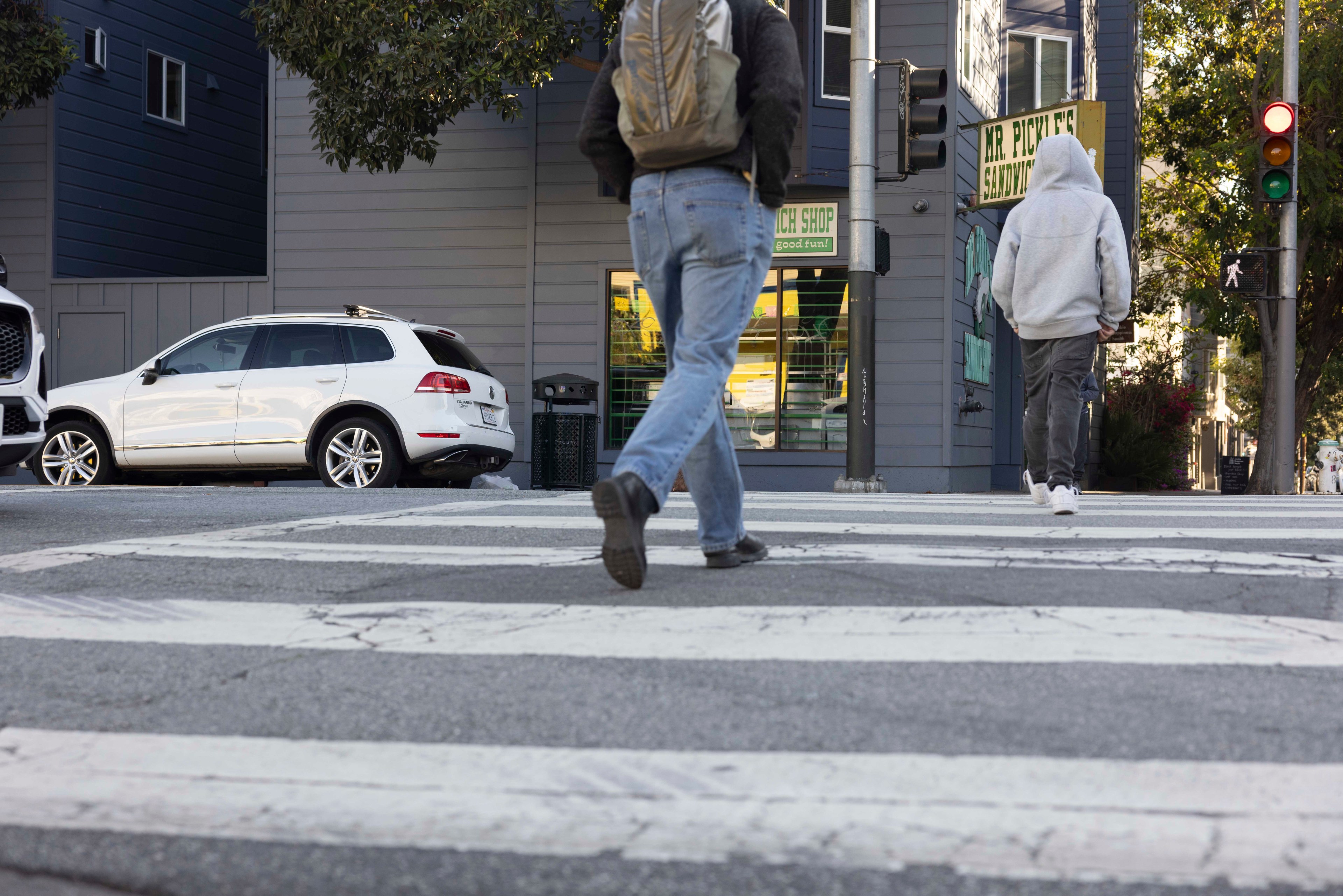 Two people cross a street with a white SUV parked nearby. One wears a backpack, and the other has a hoodie. A sign for "Mr. Pickles" is visible.