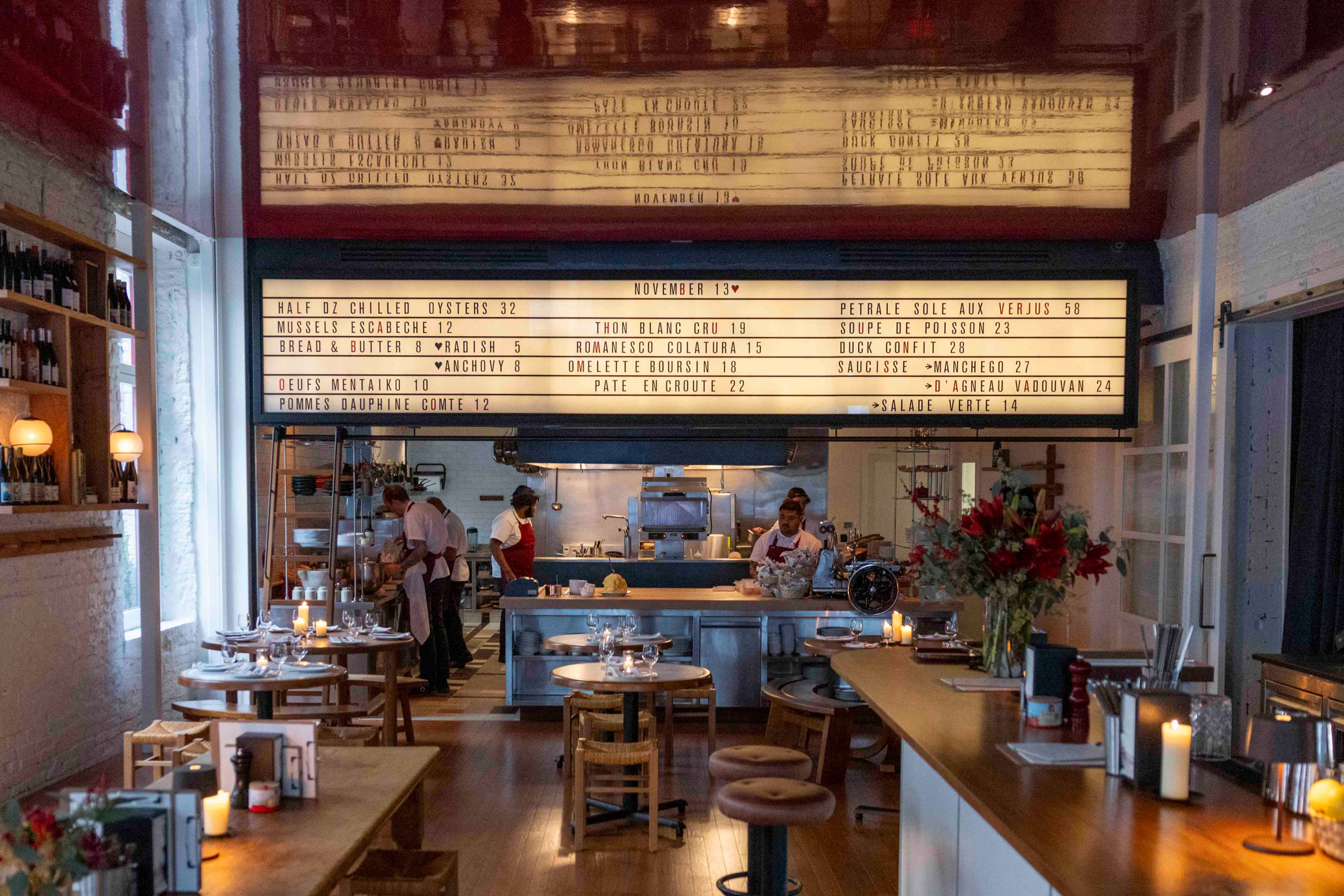 A cozy restaurant with dim lighting features a menu board above an open kitchen where chefs in aprons prepare food. Tables are set elegantly with candles.