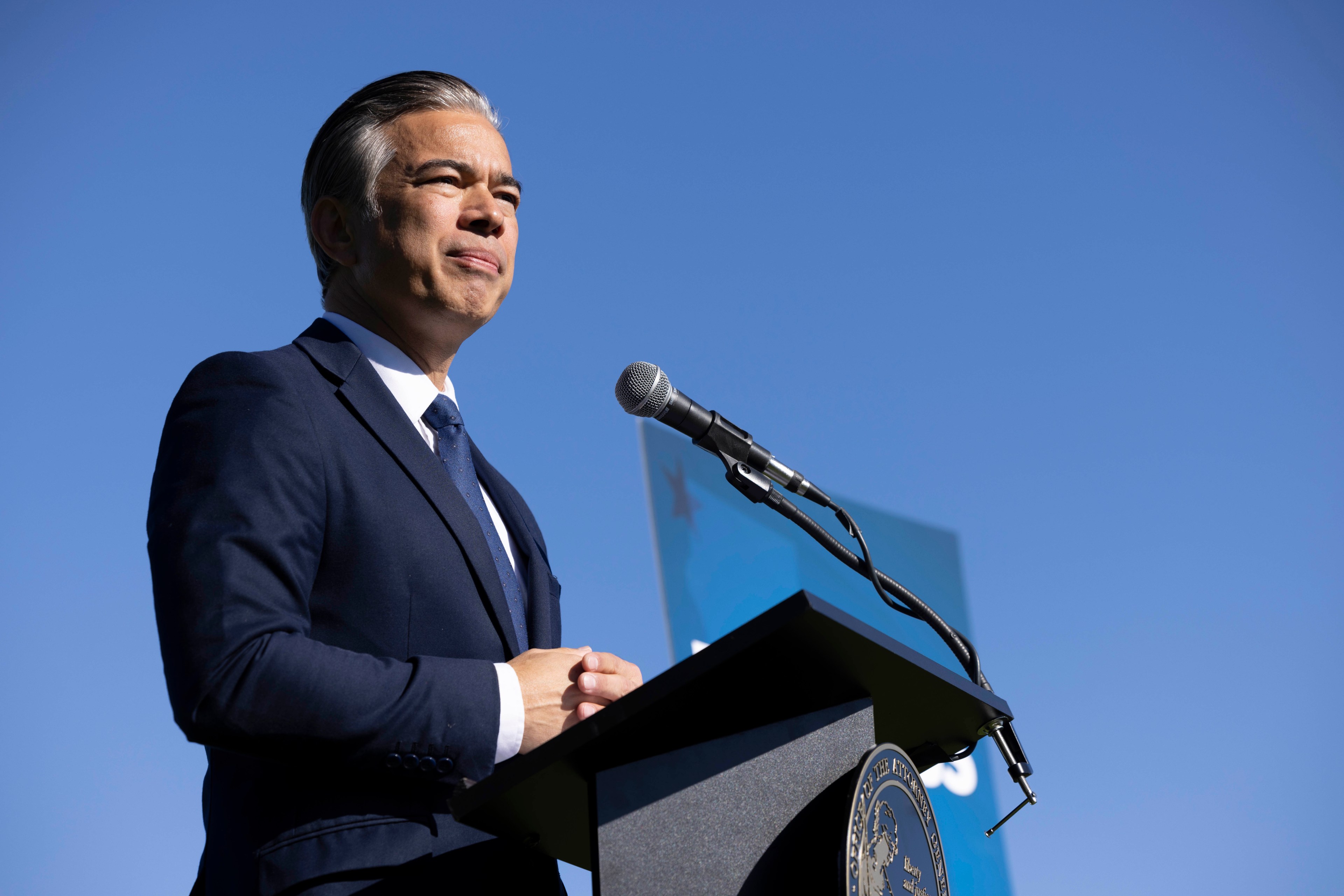 A man in a blue suit stands behind a podium with a microphone, looking thoughtful. The sky is bright blue, and a partial sign is visible in the background.