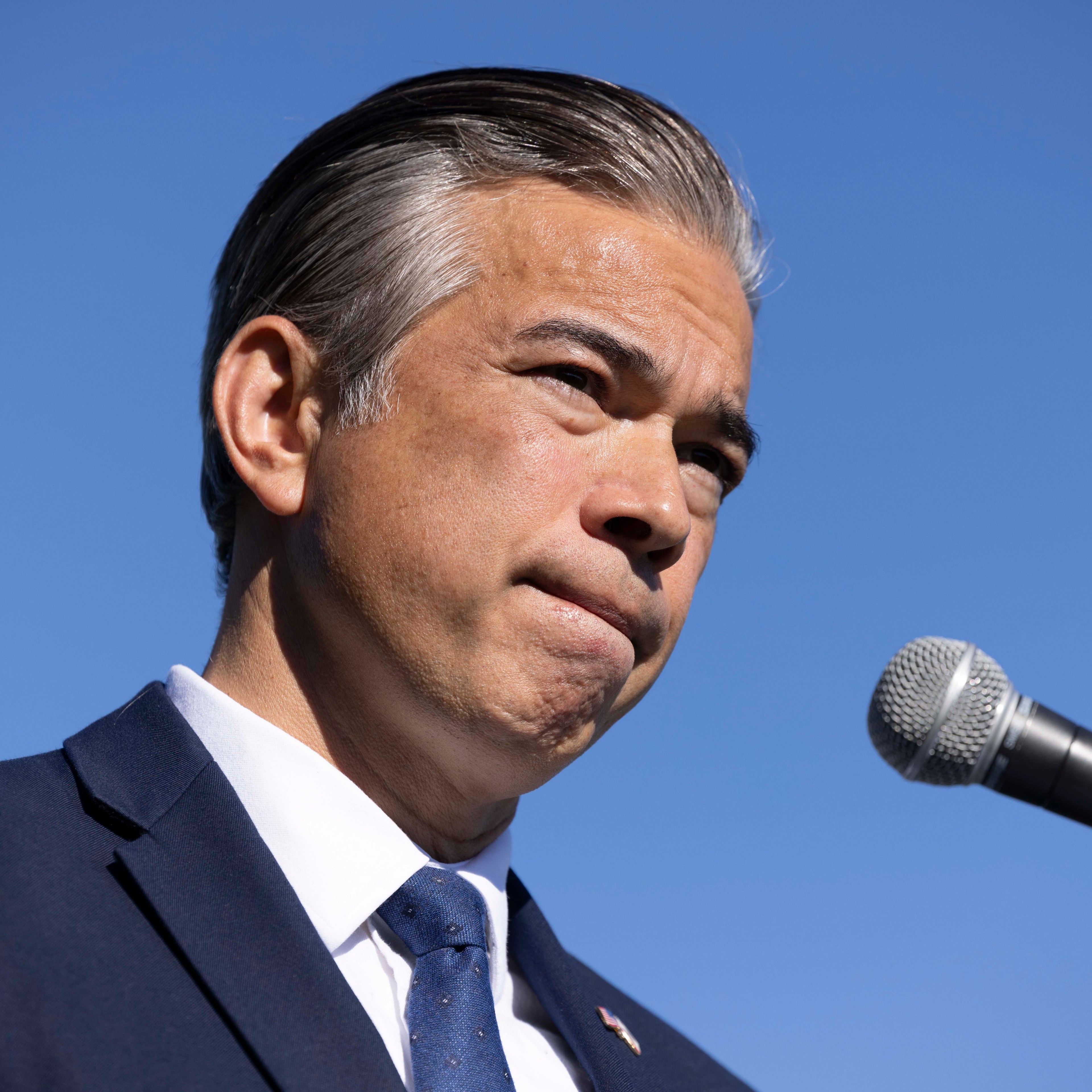 A man in a suit stands against a clear blue sky, speaking into a microphone with a focused expression.