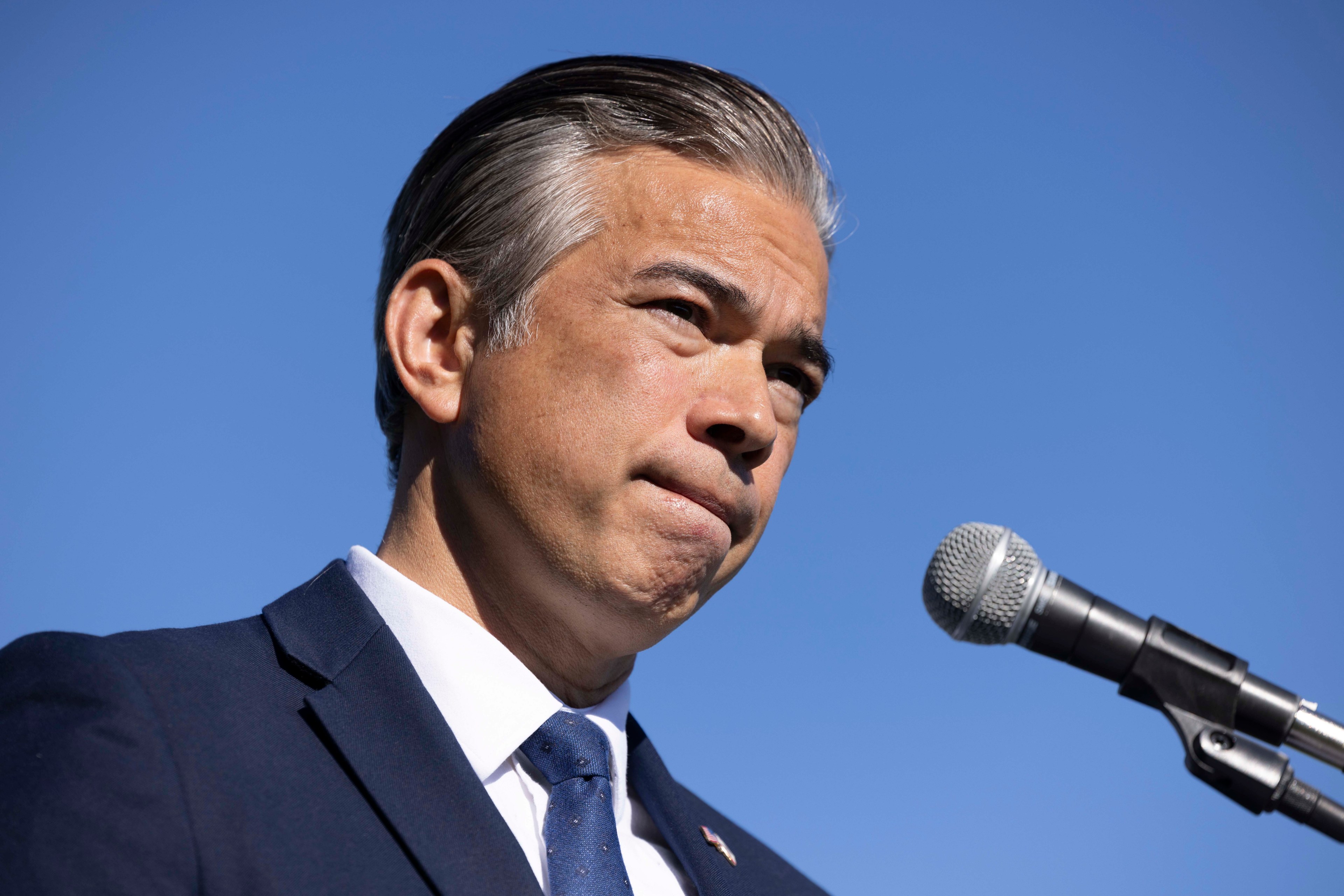 A man in a suit stands against a clear blue sky, speaking into a microphone with a focused expression.