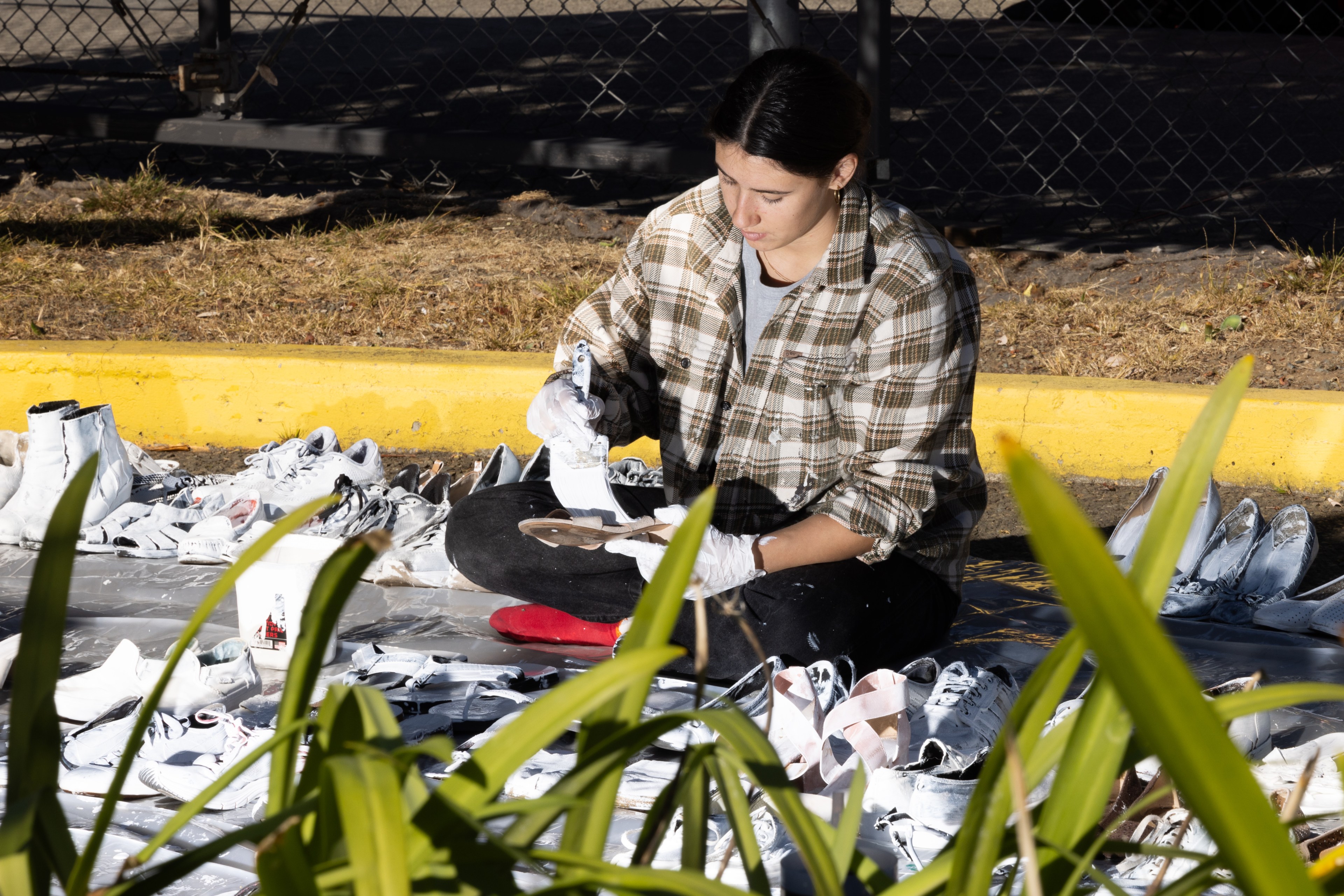 A person in a plaid shirt is sitting outdoors, surrounded by several white shoes. They appear to be painting or working on these shoes with focused attention.