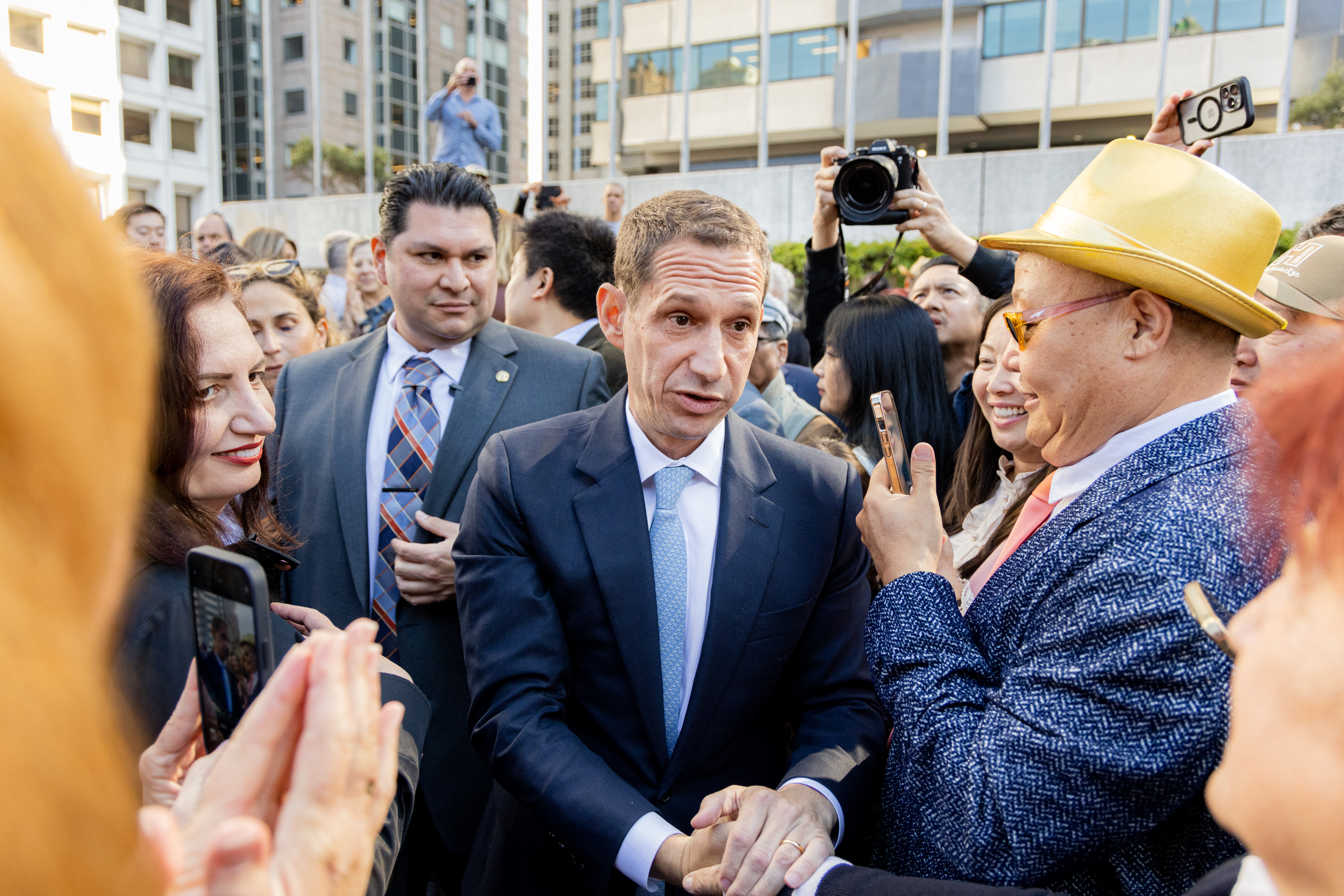 A man in a suit is shaking hands in a crowd outdoors. He is surrounded by people taking photos and smiling, with tall buildings in the background.