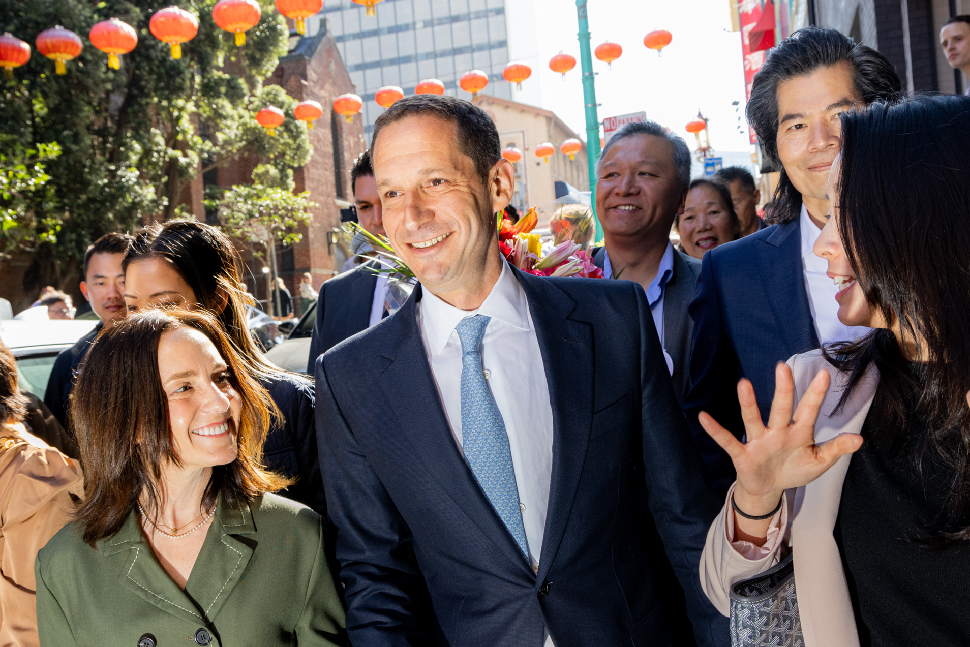 A group of people, dressed in formal and casual attire, walk down a street decorated with red lanterns, smiling and engaging with each other.