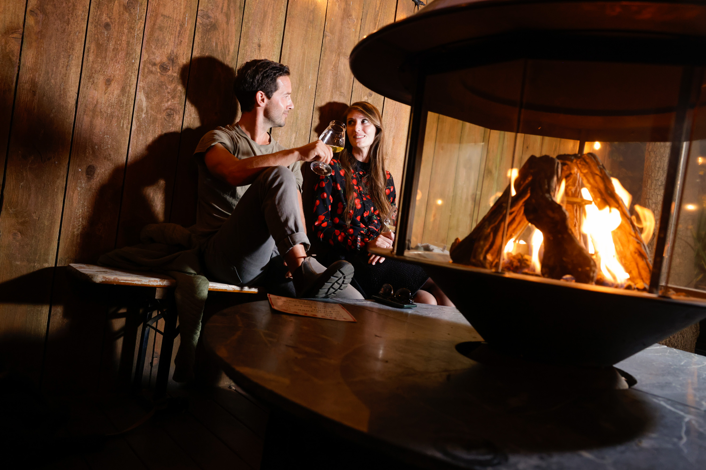 A man and woman sit on a cozy bench by a warm, glowing fireplace, holding glasses, with soft lighting and wooden walls in the background.