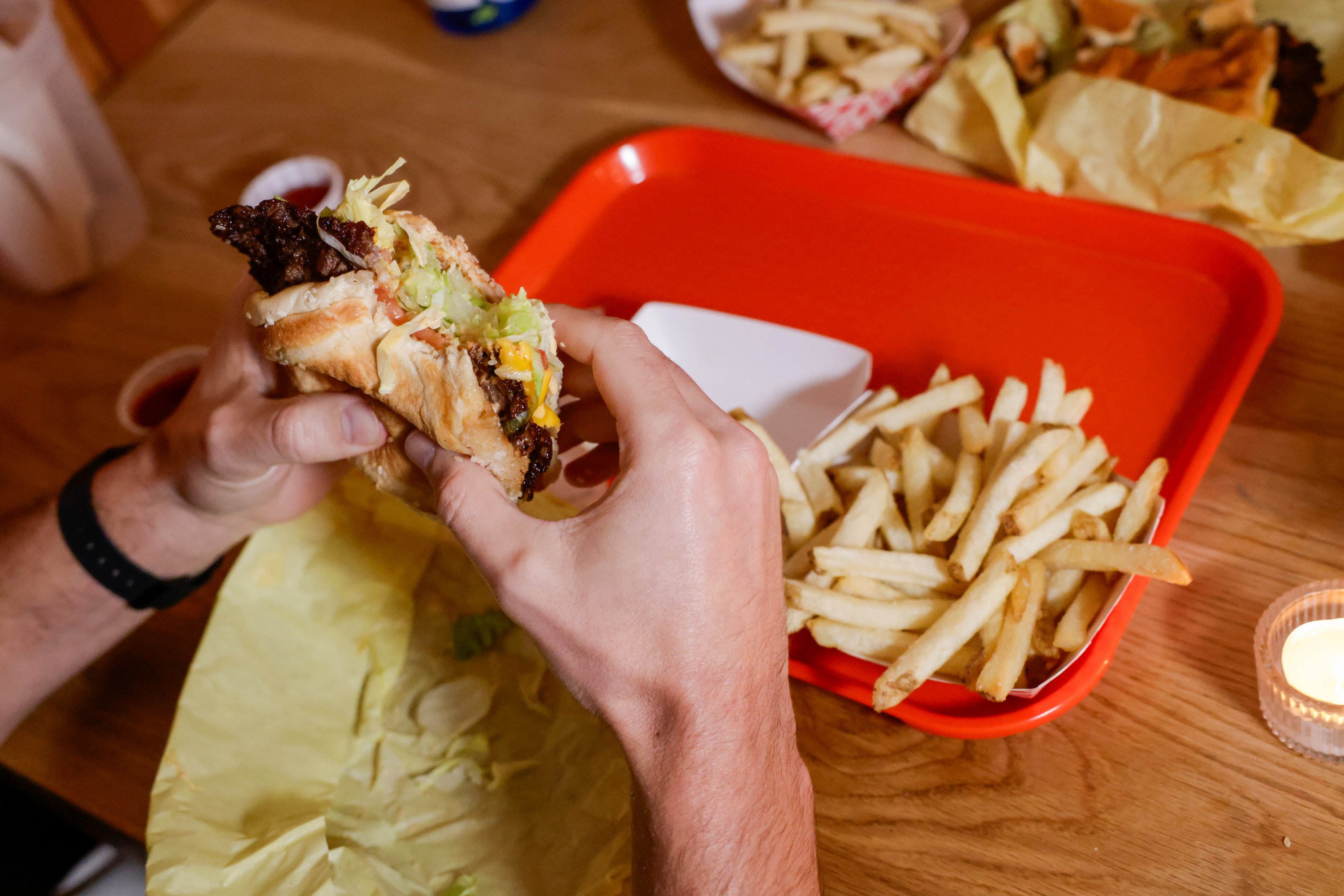 A person holds a burger over a table with a red tray. On the tray are fries, and there’s a candle nearby. Sauces appear in small cups.