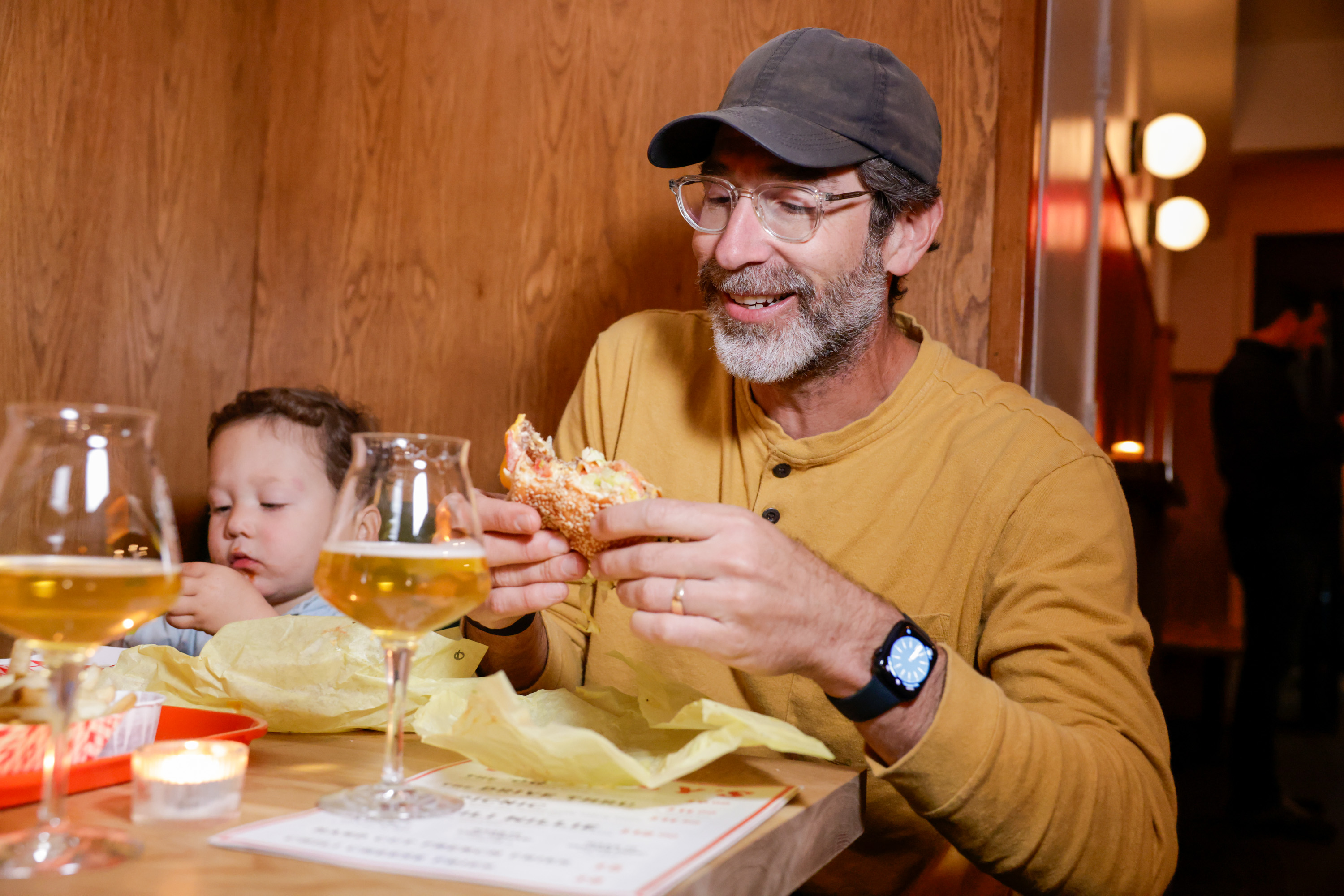 A man in a yellow shirt and cap enjoys a sandwich, sitting at a table with a child. There are two glasses of beer, a candle, and food in yellow wrappers.